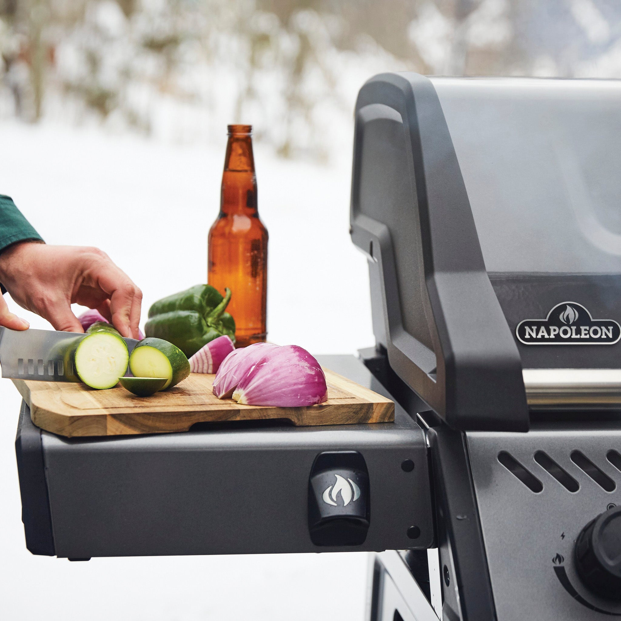 A person slicing vegetables next to a Napoleon Freestyle 365 Gas Barbecue with cast iron cooking grids and a beer bottle placed on the side shelf.