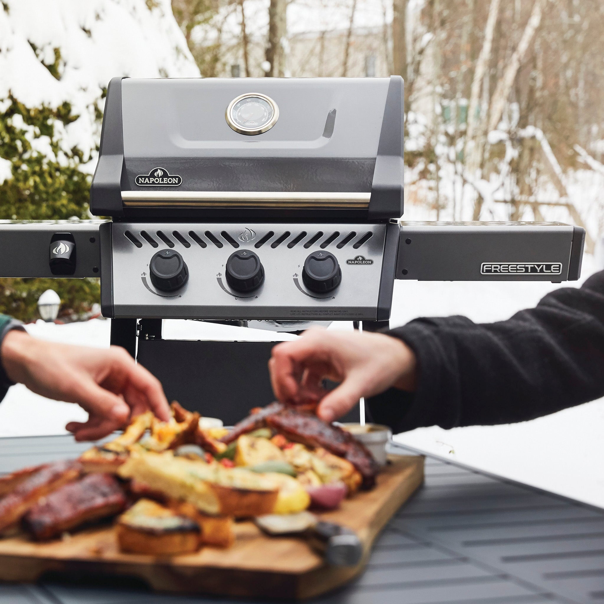 Two people enjoy food from a Napoleon Freestyle 365 Gas Barbecue equipped with cast iron cooking grids, with a wooden board of perfectly cooked meats and vegetables in front.