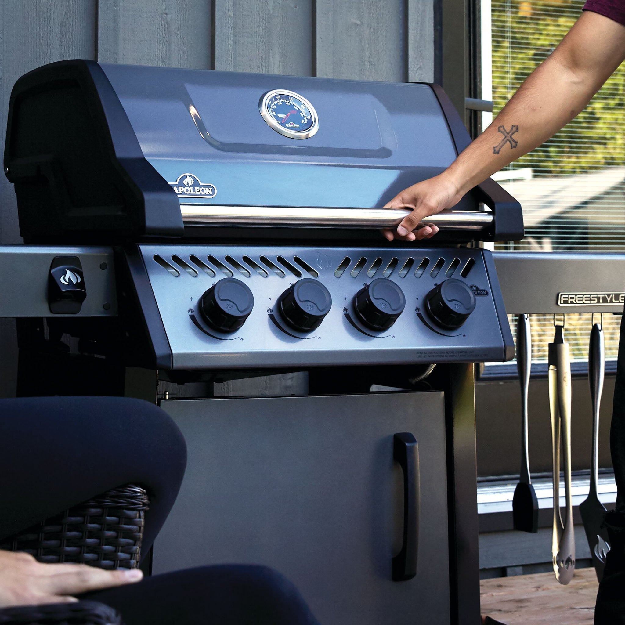 A person opening a Napoleon Freestyle 425 SIB Gas Barbecue with the JETFIRE ignition system on a patio, with grill utensils hanging on a side shelf.