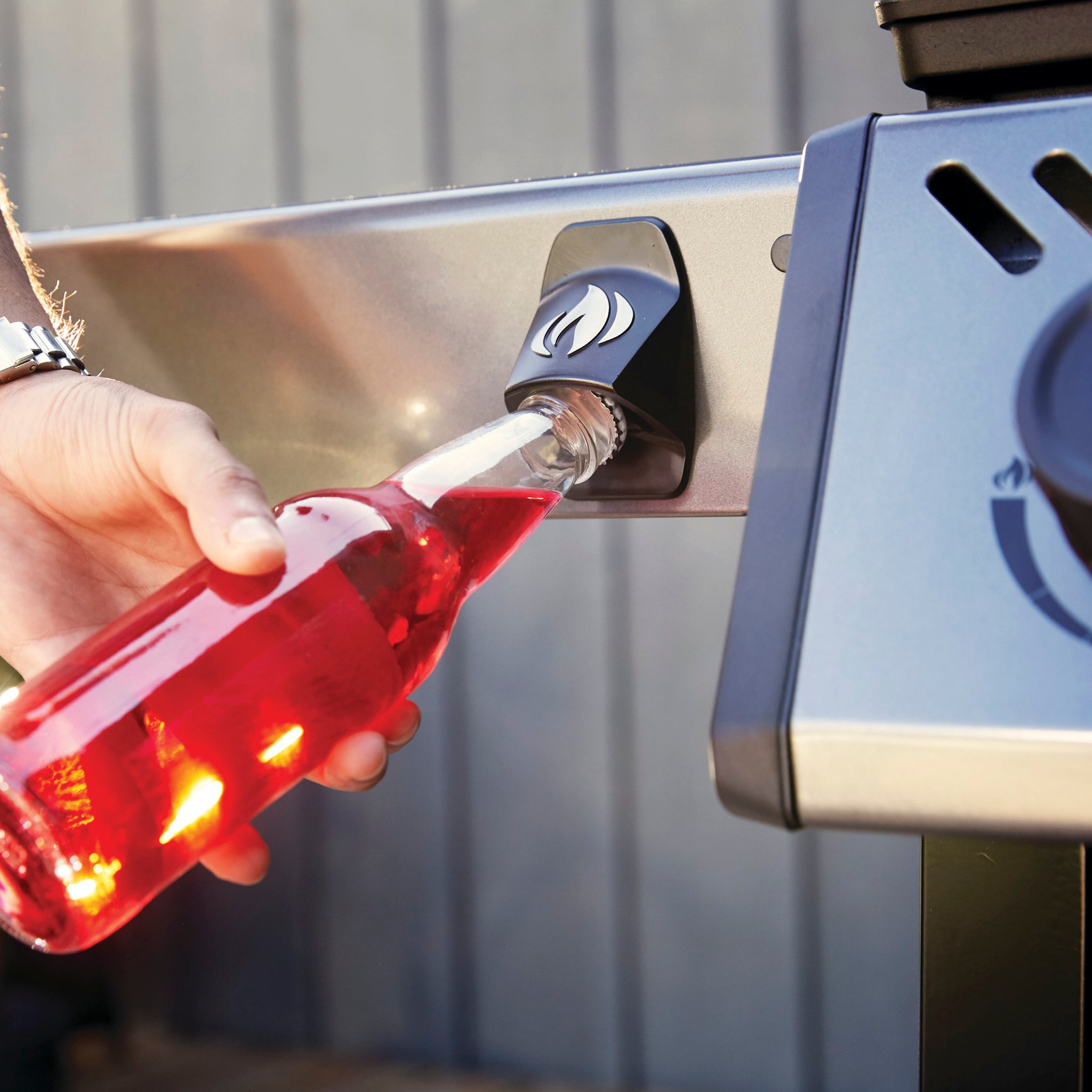 Person opening a bottle of red beverage using a built-in bottle opener on their Napoleon Freestyle 425 Gas Barbecue, set beside cast iron porcelainized enamel cooking grids and the sleek JETFIRE ignition system.
