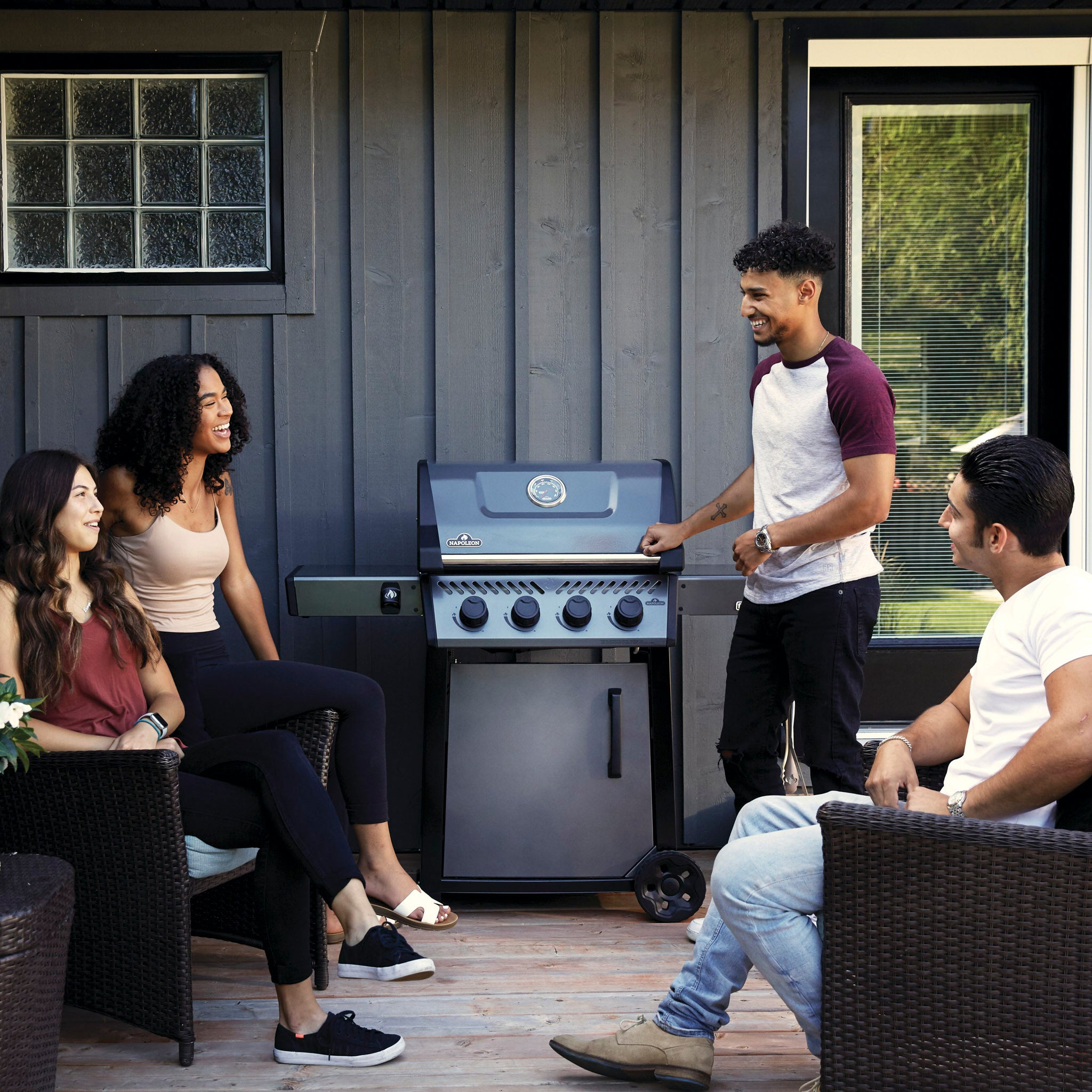 Four friends enjoying a conversation while standing and sitting around a Napoleon Freestyle 425 Gas Barbecue with cast iron porcelainized enamel cooking grids on a wooden deck.