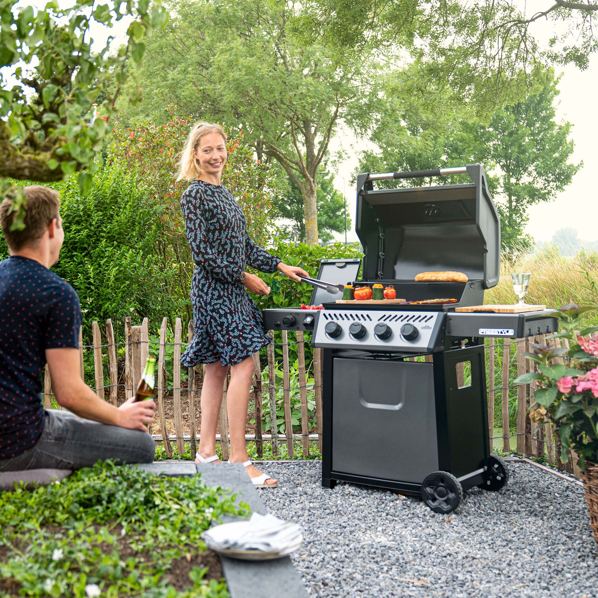 In a garden setting, a woman grills food outdoors on a Napoleon Freestyle 425 SIB Gas Barbecue, featuring the efficient JETFIRE ignition system, as a man sits nearby holding a drink.