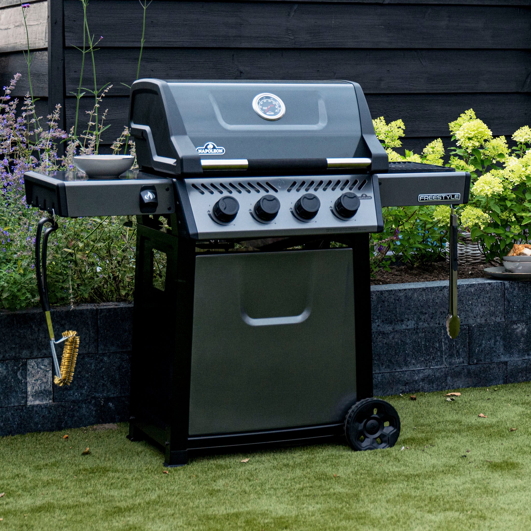 A Napoleon Freestyle 425 gas barbecue, black in color, featuring four knobs, side burners, and grilling tools along with a JETFIRE ignition system and cast iron porcelainized enamel cooking grids, is set on a lawn with flowers in the background.