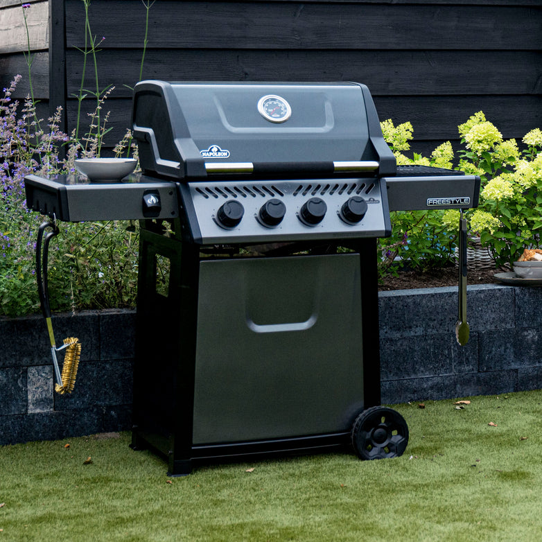 A Napoleon Freestyle 425 gas barbecue, black in color, featuring four knobs, side burners, and grilling tools along with a JETFIRE ignition system and cast iron porcelainized enamel cooking grids, is set on a lawn with flowers in the background.