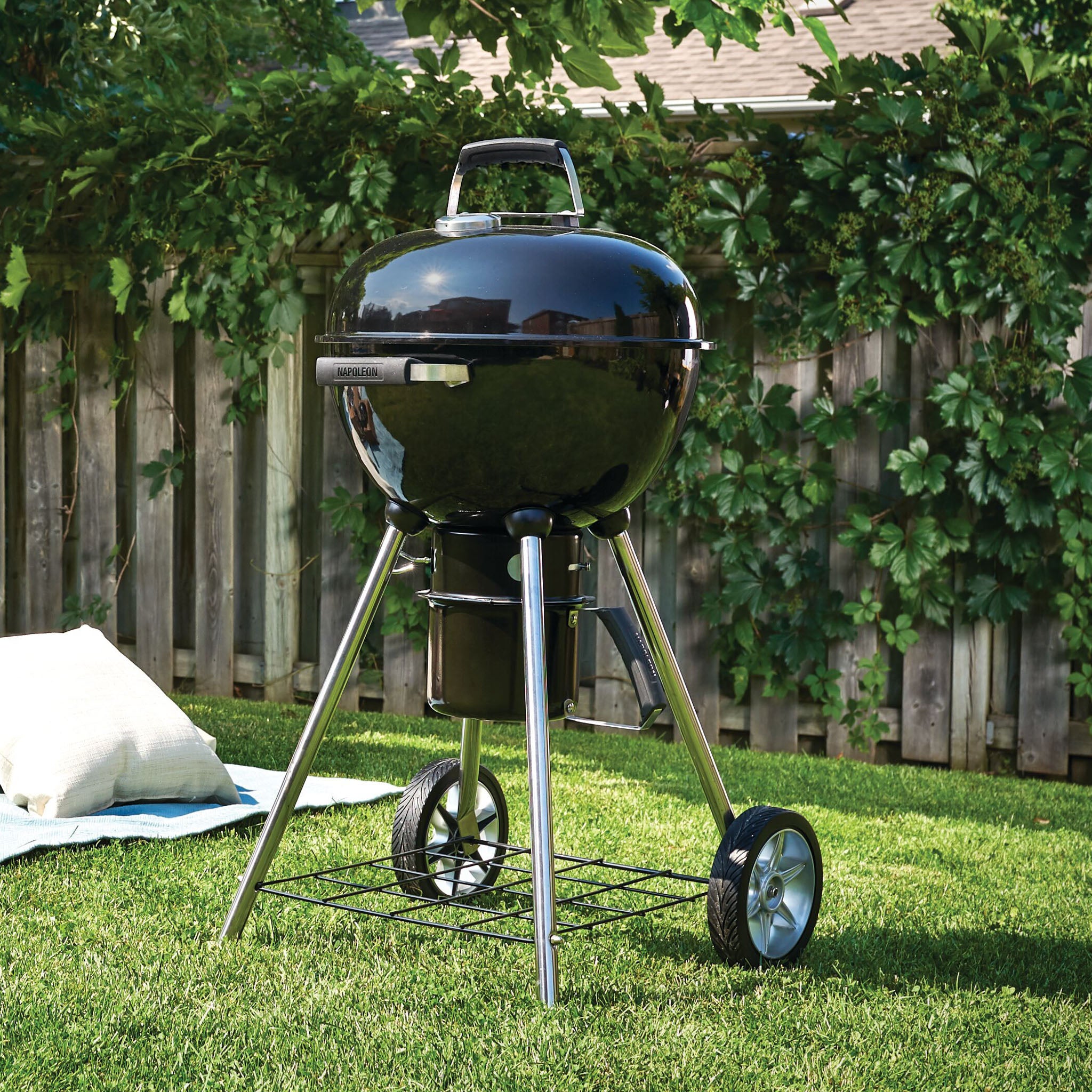 The Napoleon NK18 Charcoal Kettle Grill, with its adjustable vents, is set on a green lawn near a white cushion and wooden fence, perfectly complemented by the greenery in the background.