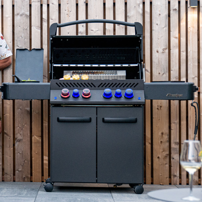 Outdoor grill with lid open, showcasing the Napoleon Phantom Prestige® 500 RSIB Gas Barbecue's infrared burners, encased by a wooden fence. A wine glass is placed on the right, and a person is partially visible on the left. The stainless steel cooking grids ensure even heat distribution for perfect grilling every time.