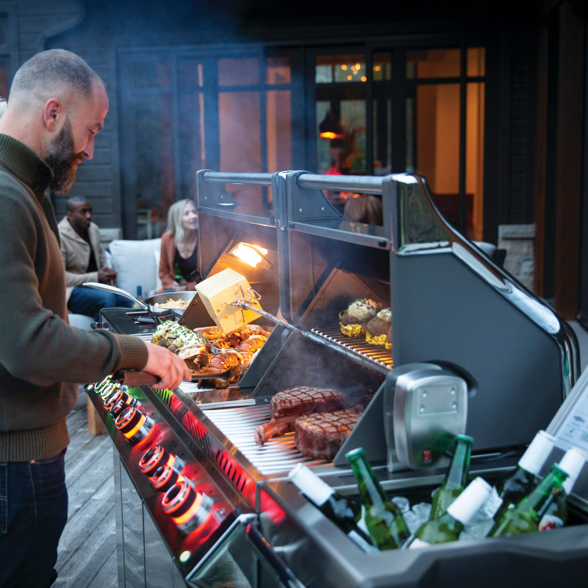 A man expertly grills food on his Napoleon Prestige Pro 825 RSIB Gas Barbecue, while people chat in the background near the outdoor seating area. The grill's impressive infrared burners ensure perfectly cooked meals.