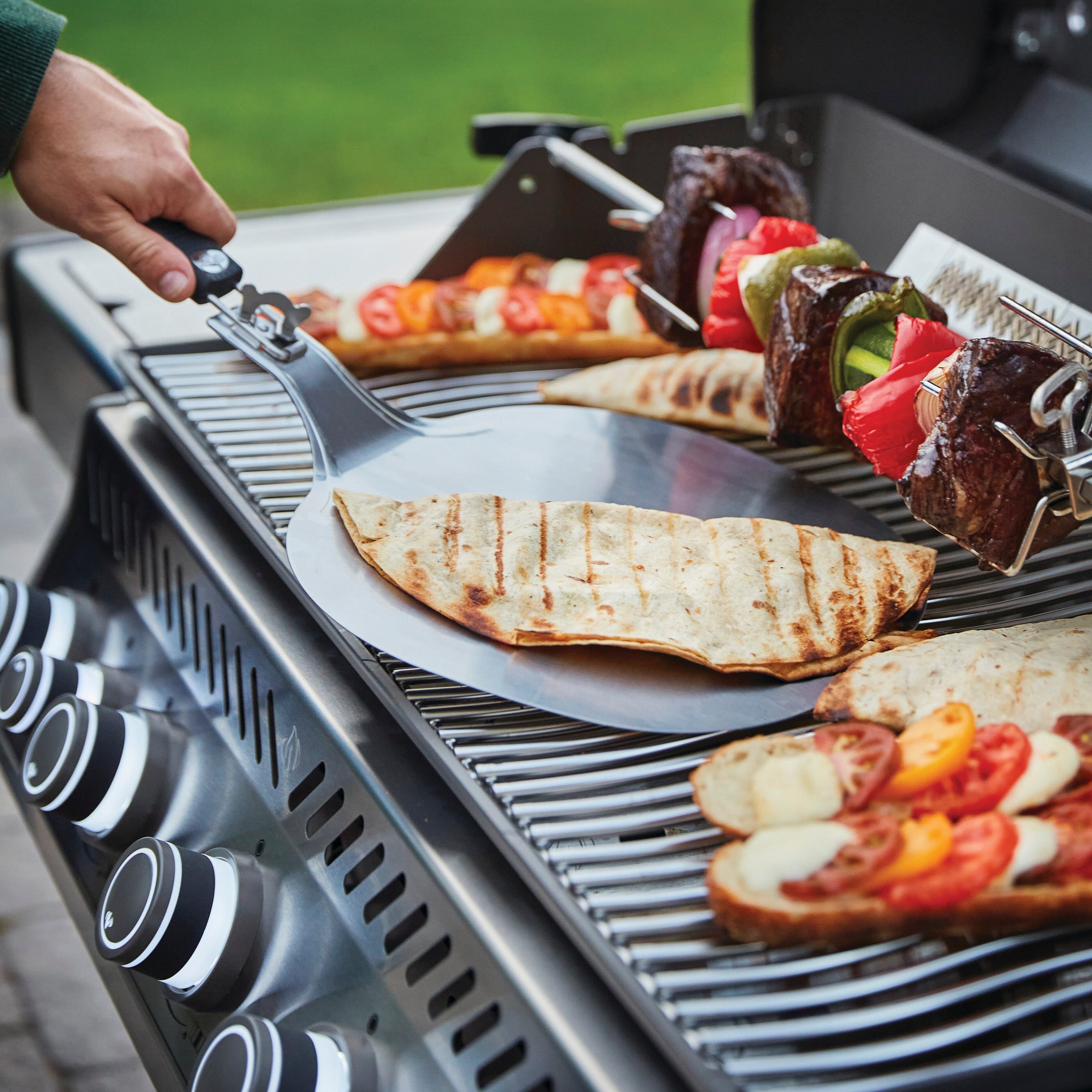 A person grilling meat and vegetables on a Napoleon Rogue SE 625 RSIB Gas Barbecue with flatbreads on a metal spatula in front, outdoors.