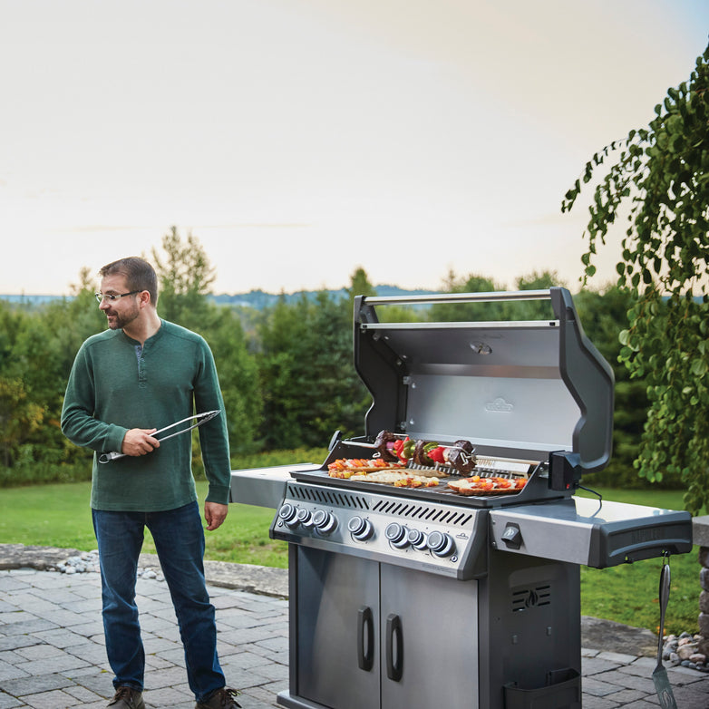 A man in a green sweater grills skewers on a Napoleon Rogue SE 625 RSIB Gas Barbecue with an infrared side burner, set in a backyard with lush greenery.