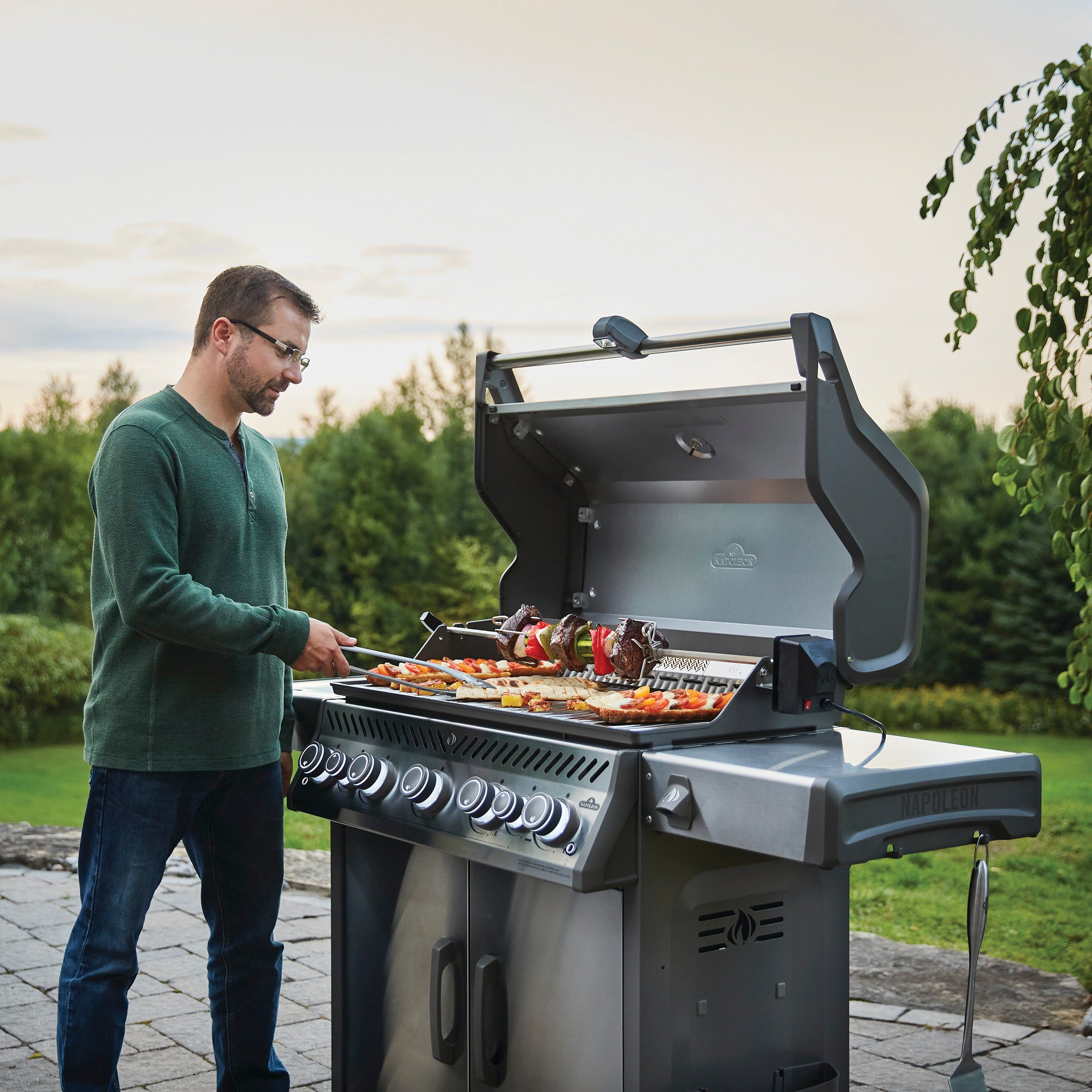 A man grills skewers and vegetables on the Napoleon Rogue SE 625 RSIB Gas Barbecue with its efficient infrared side burner, in a scenic backyard setting.