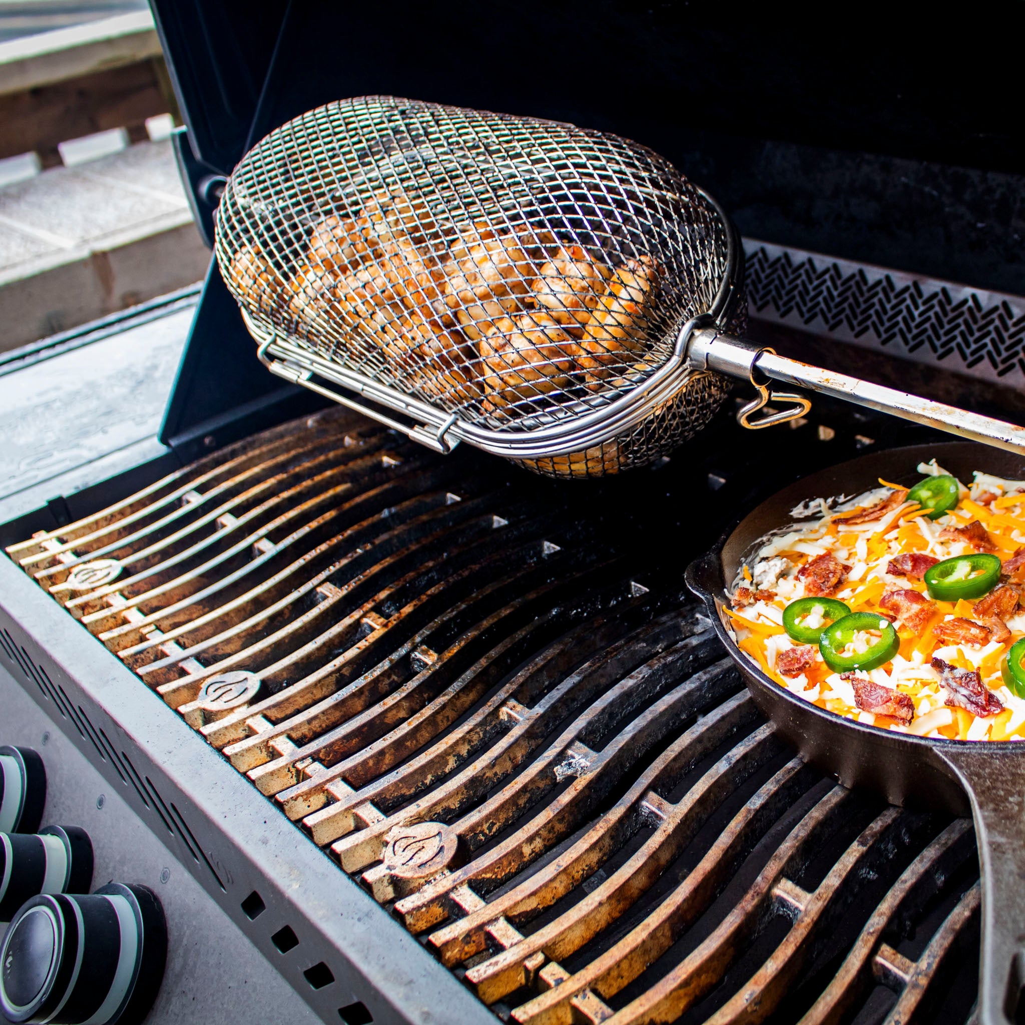 Chicken drumsticks cooking in a Napoleon Stainless Steel Rotisserie Basket on the grill, with a skillet of cheesy jalapeño dish beside it.