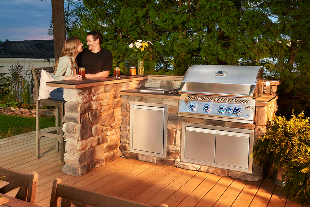 A couple relaxes at an outdoor stone kitchen bar featuring the Burford Grand Sideboard with Wine Rack in Navy Blue, surrounded by trees as evening light filters through.