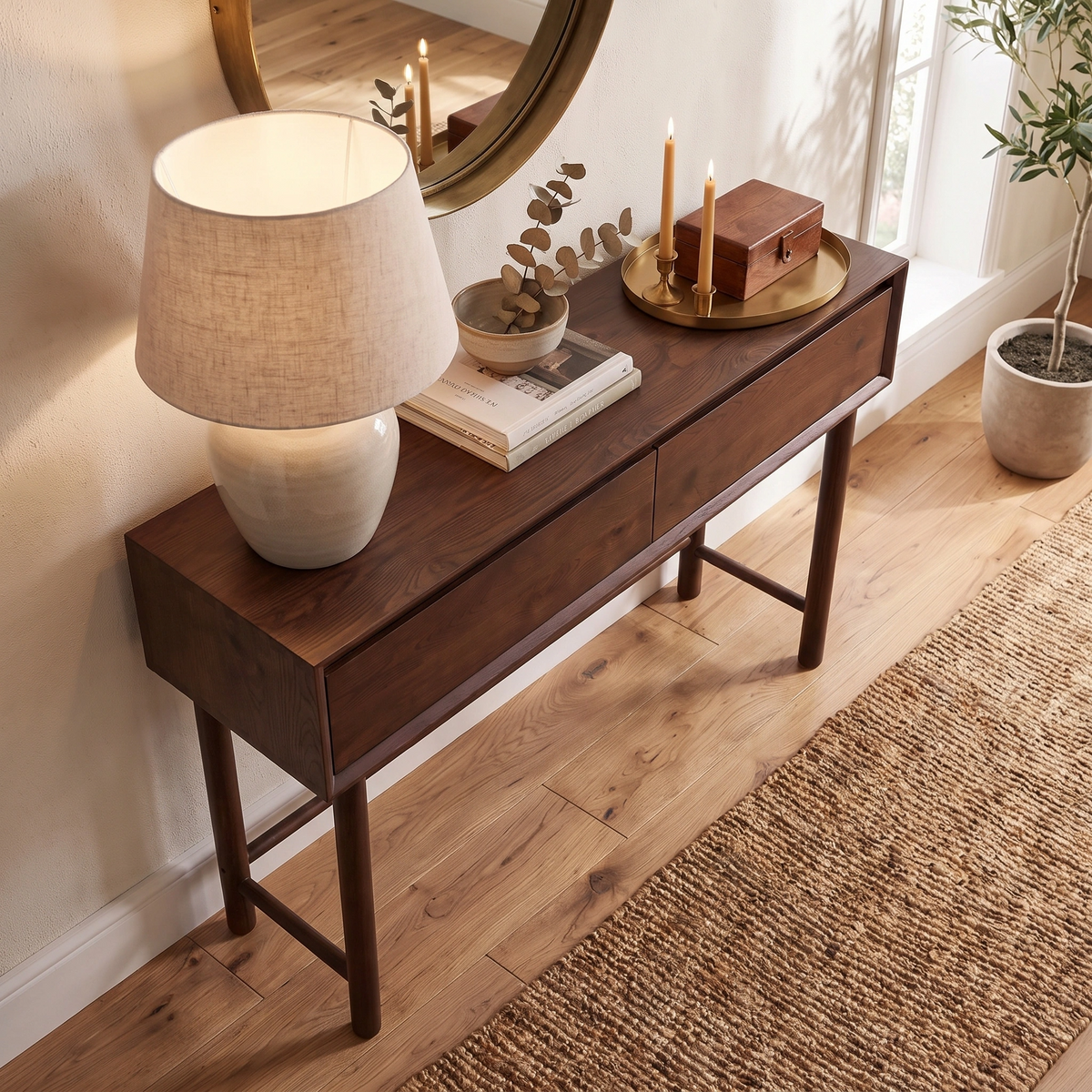 The Oslo 2 Drawer Console Table in Walnut is adorned with a lamp, books, candles, and a tray, and is positioned on a wooden floor near a window and mirror.
