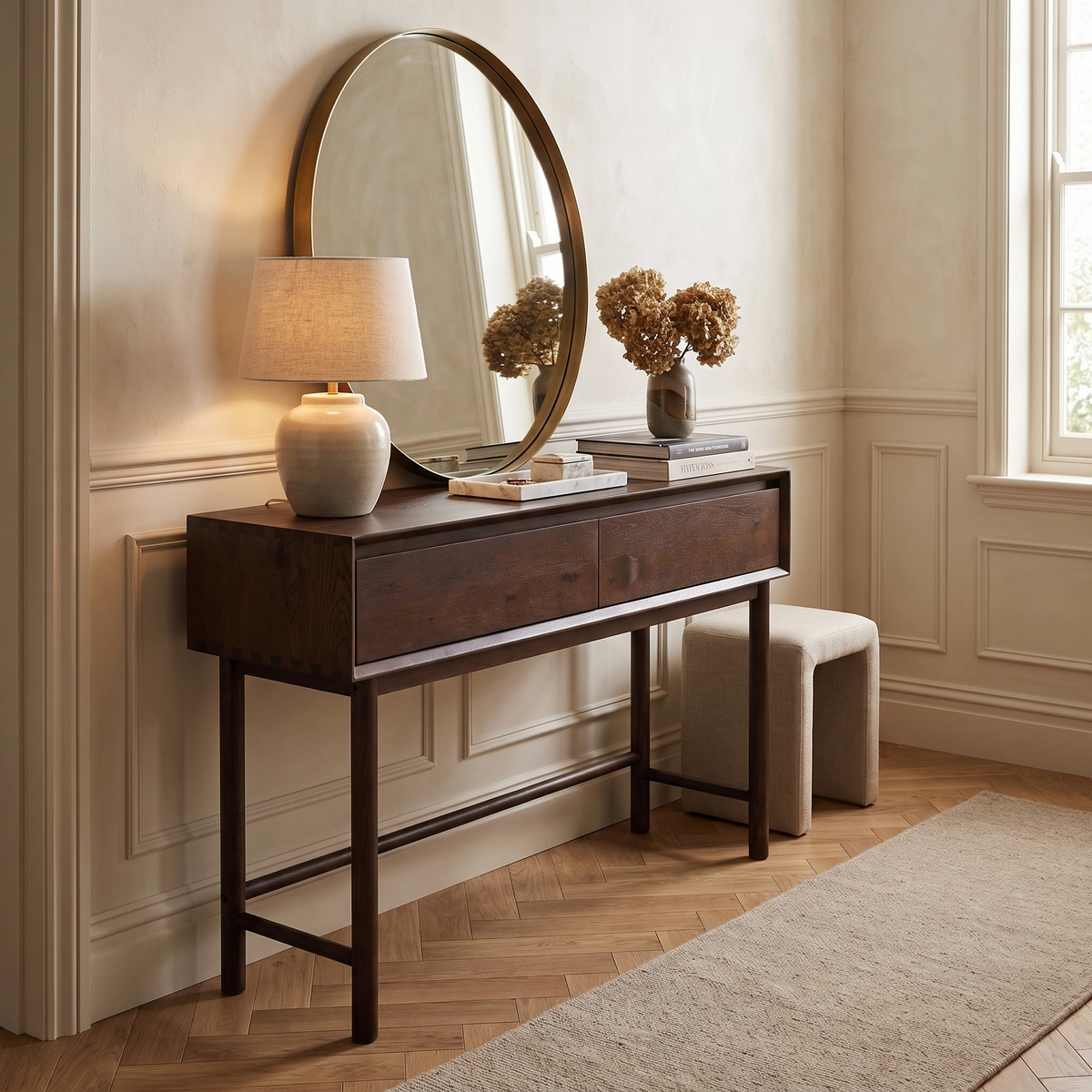 A walnut Oslo 2 Drawer Console Table with a round mirror, lamp, books, and dried flowers is placed beside a window in a bright room.