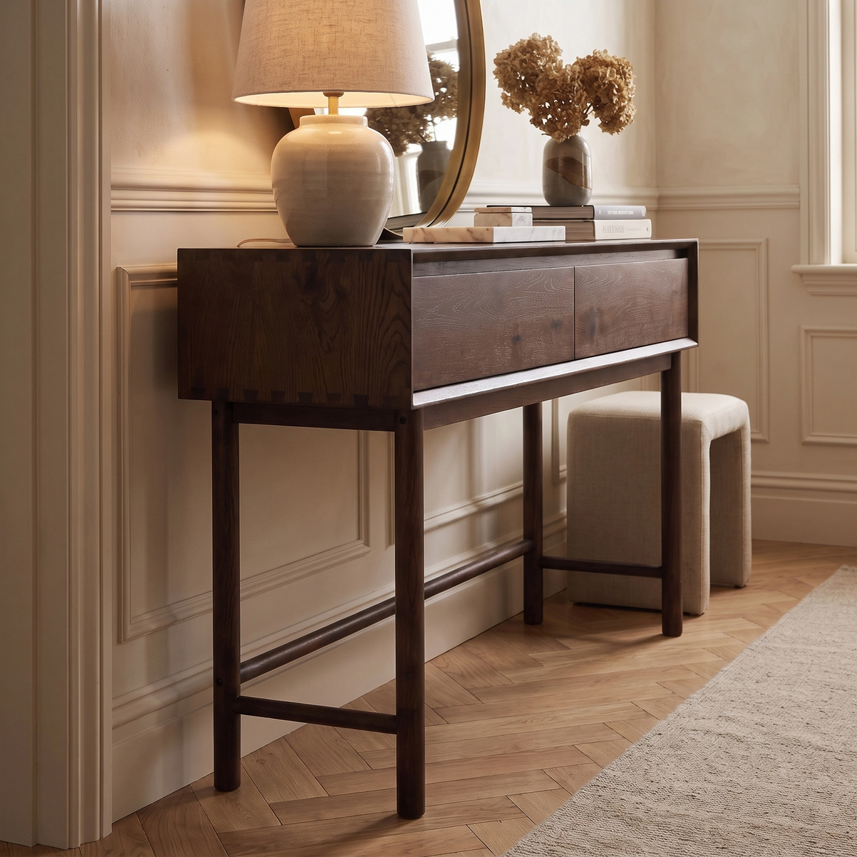 An Oslo 2 Drawer Console Table in Walnut, adorned with a lamp, books, and dried flowers, sits against a beige paneled wall.