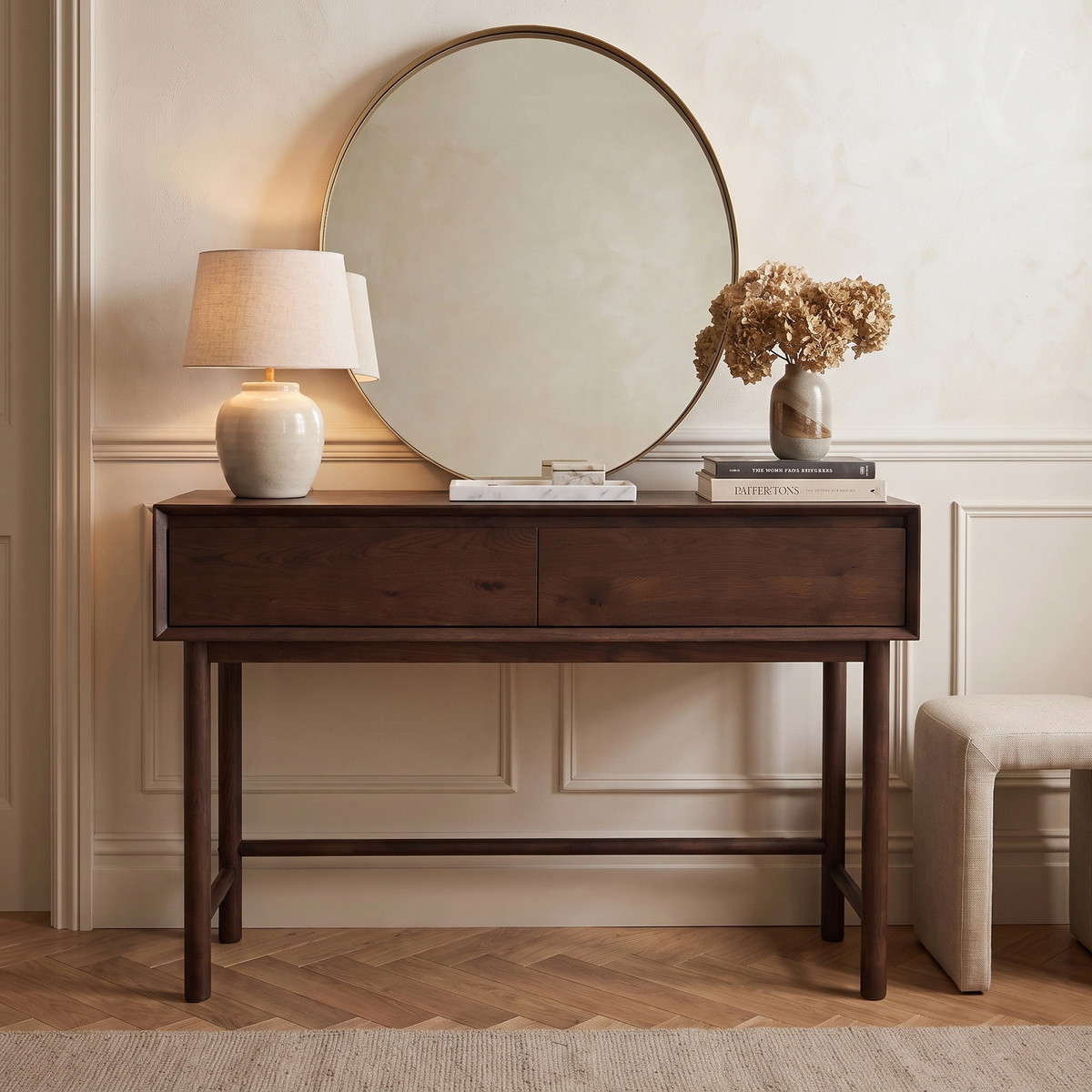 An Oslo 2 Drawer Console Table in Walnut styled with a lamp, books, a vase of dried flowers, and topped with a round mirror above.