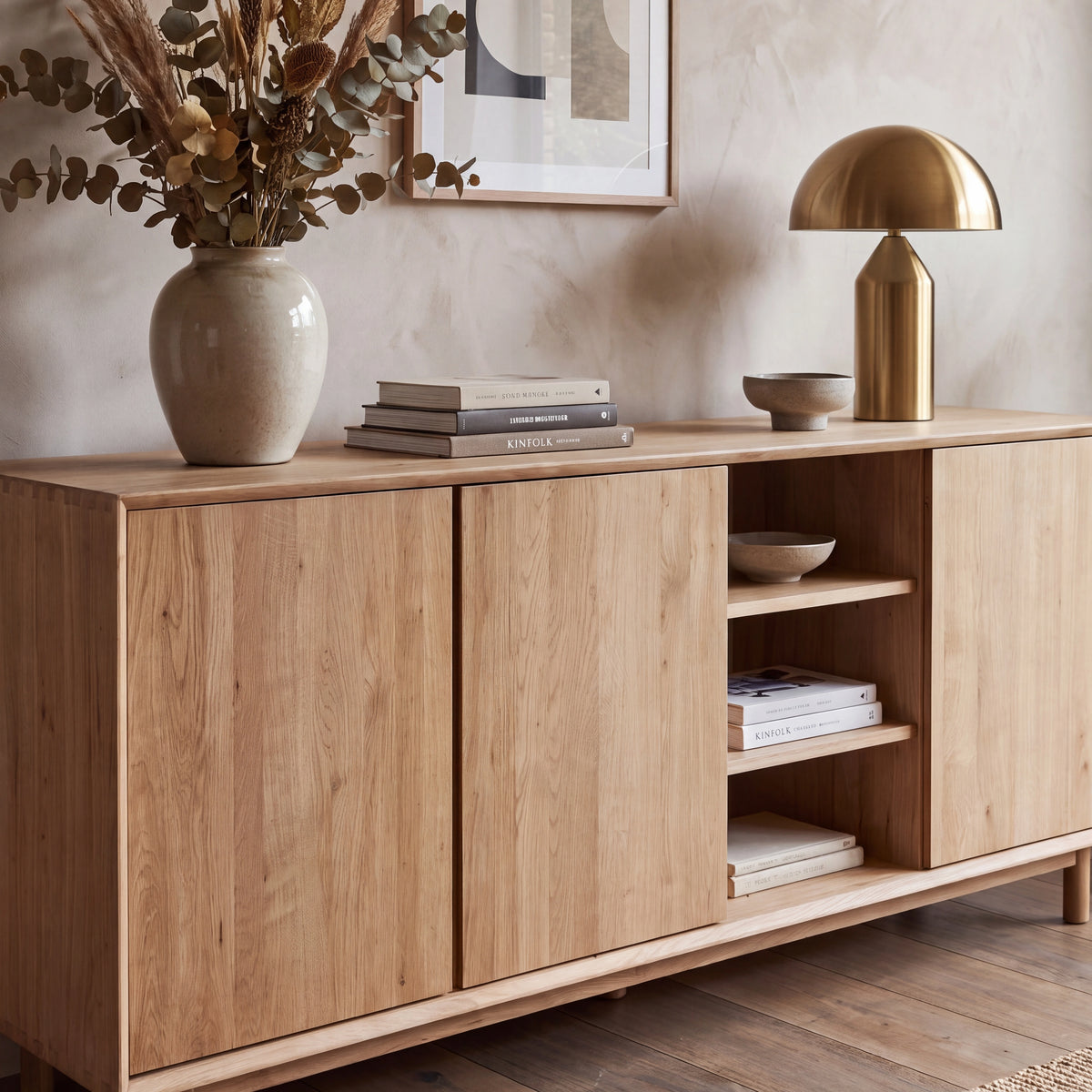 The Oslo 3 Door Sideboard with Shelves in Light Oak is styled with books, a vase of dried flowers, a gold lamp, and decorative bowls on top. It features adjustable shelving to suit your storage needs while maintaining a stylish and contemporary look.
