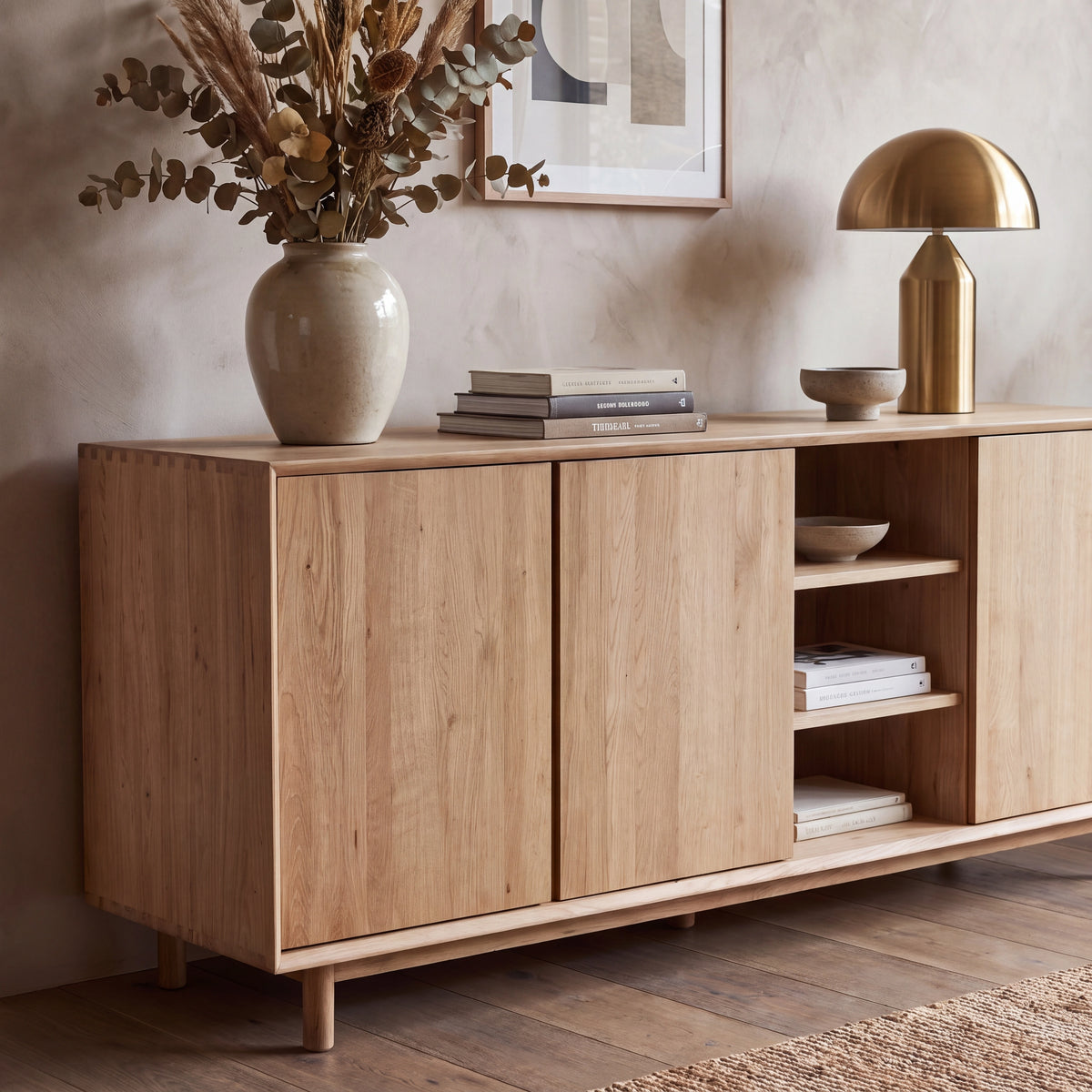 The Oslo 3 Door Sideboard with Shelves in Light Oak, featuring adjustable shelving, is styled with books, a ceramic vase of dried flowers, and a gold lamp placed on top.