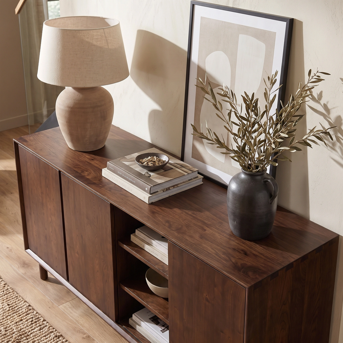 The Oslo 3 Door Sideboard with Shelves in Walnut is styled with a lamp, framed art, books, a black vase filled with greenery, and a small tray.