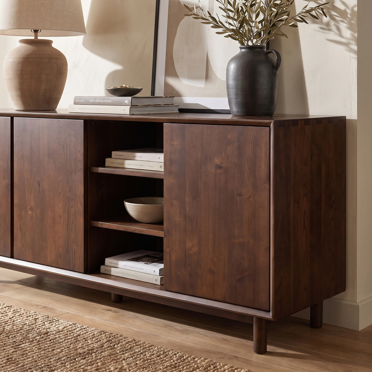 A Oslo 3 Door Sideboard with Shelves in Walnut, styled with books, vases, and framed art on and above it, sits beside a woven rug.