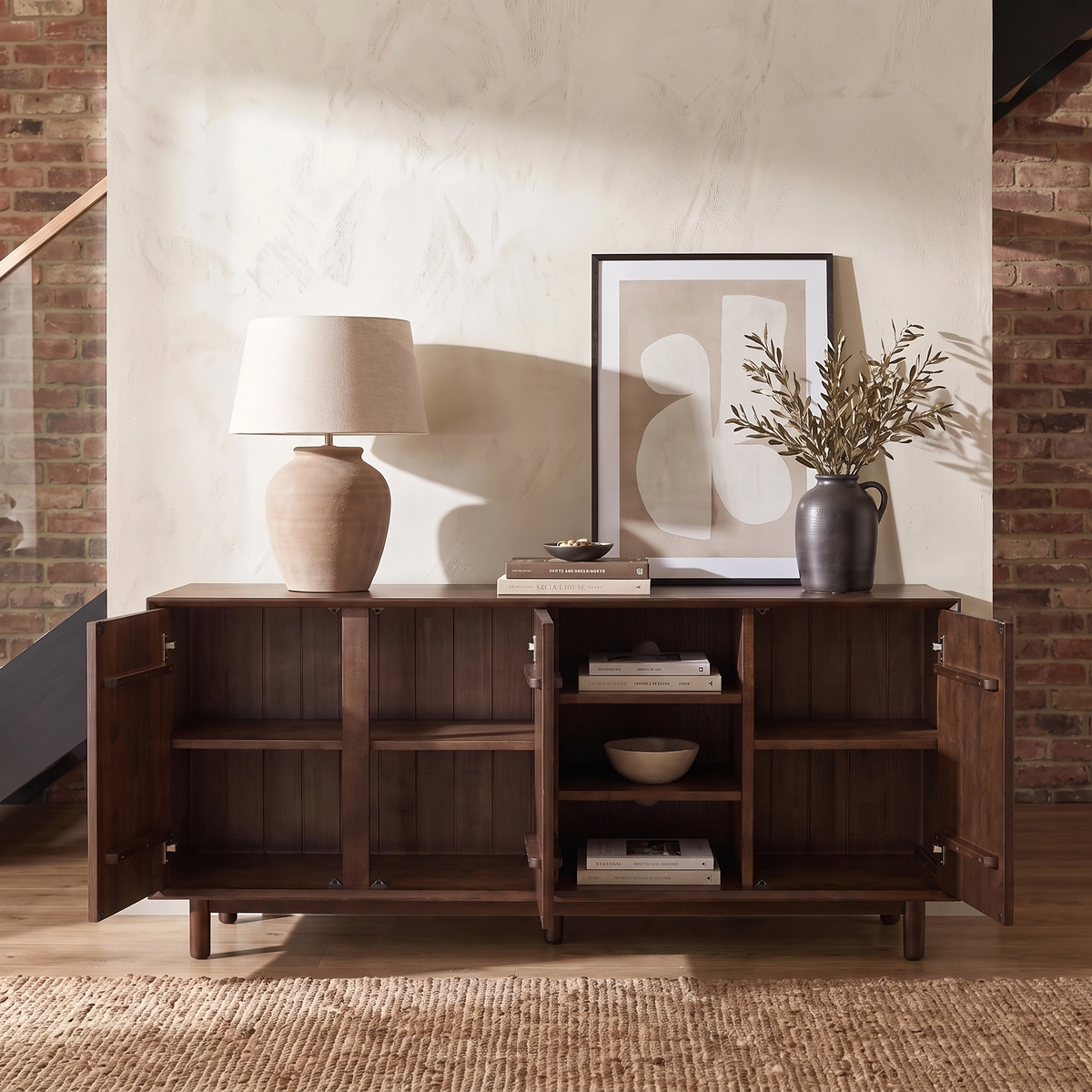 With its open doors revealing generous shelving, the Oslo 3 Door Sideboard with Shelves in Walnut provides ample storage, while a lamp, framed art, vase with branches, and pottery are elegantly arranged on top. This piece seamlessly blends modern style and practical functionality for any contemporary space.
