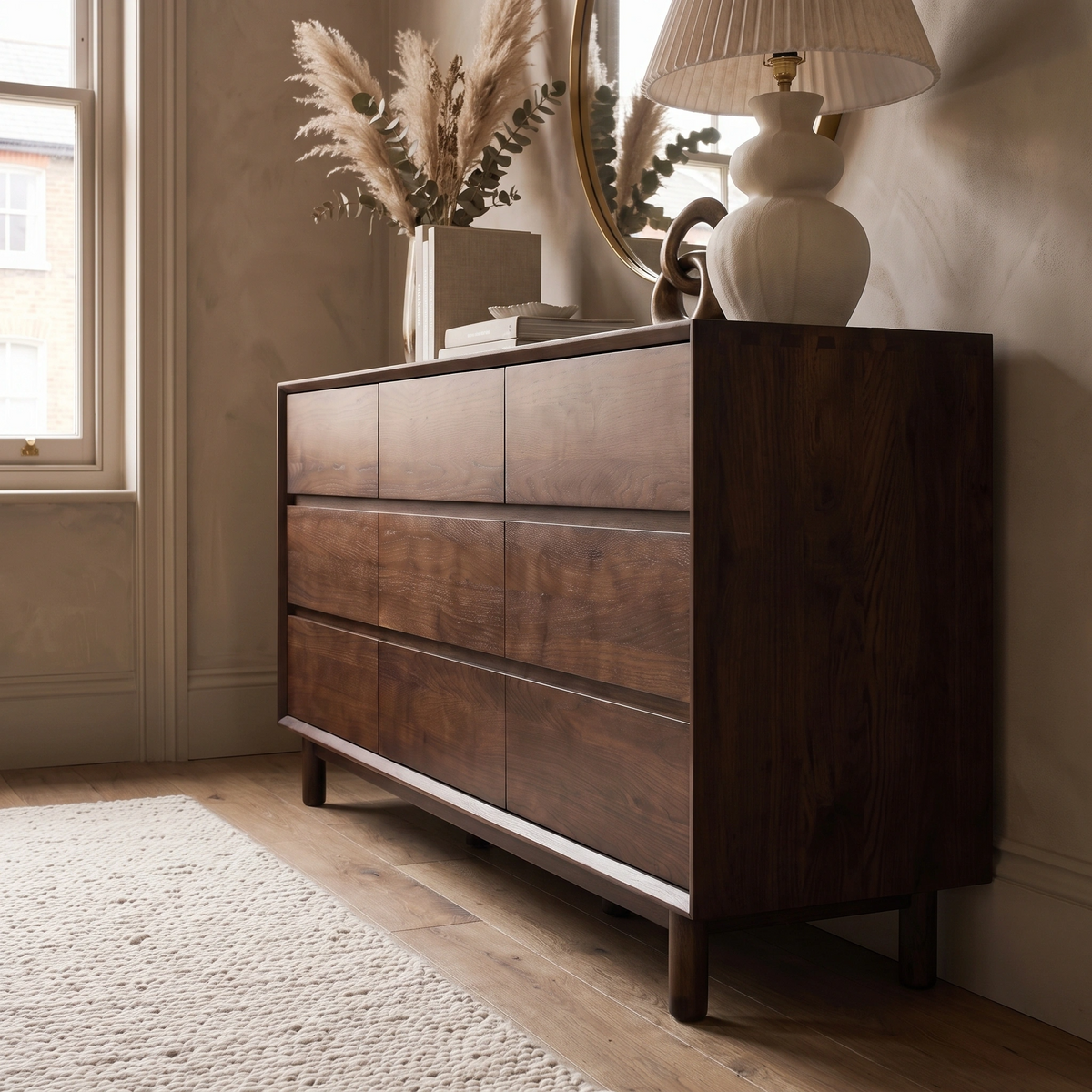 A dark wood dresser, such as the Oslo 9 Drawer Chest in Walnut, with a lamp, books, and pampas grass decor stands in a sunlit, modern room—its rich walnut finish beautifully complementing the elegance of solid oak bedroom furniture.