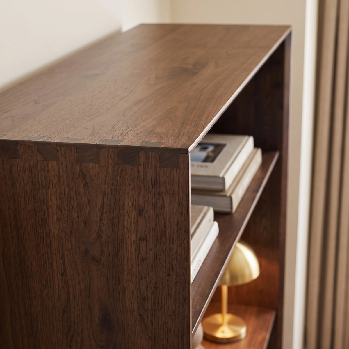 An Oslo Bookcase in Walnut, styled with books and a small gold lamp on a lower shelf, positioned beside beige curtains.