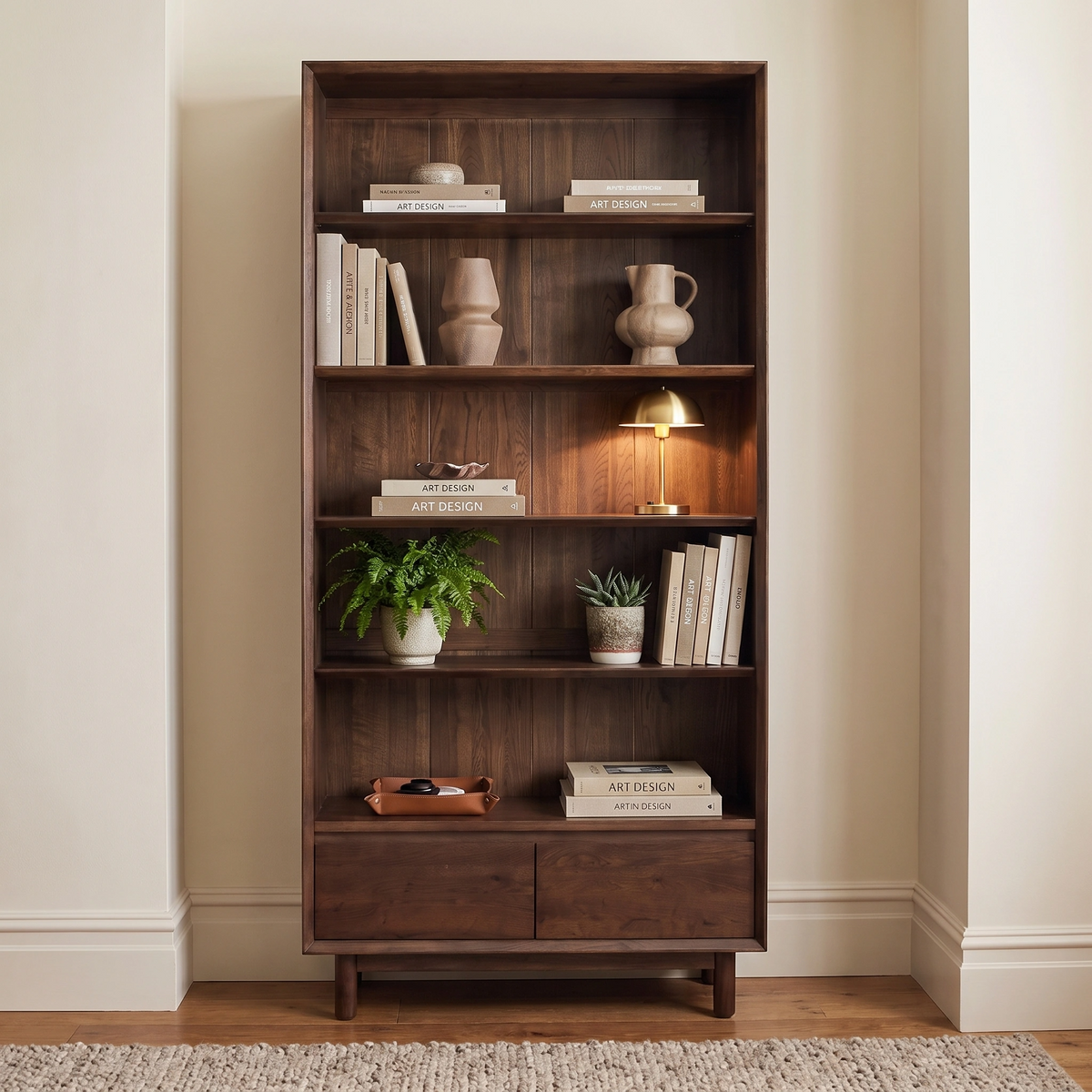 An elegant Oslo Bookcase in Walnut serves as a wooden bookshelf, beautifully displaying books, two plants, ceramic vases, a small lamp, and featuring two spacious drawers at the bottom.