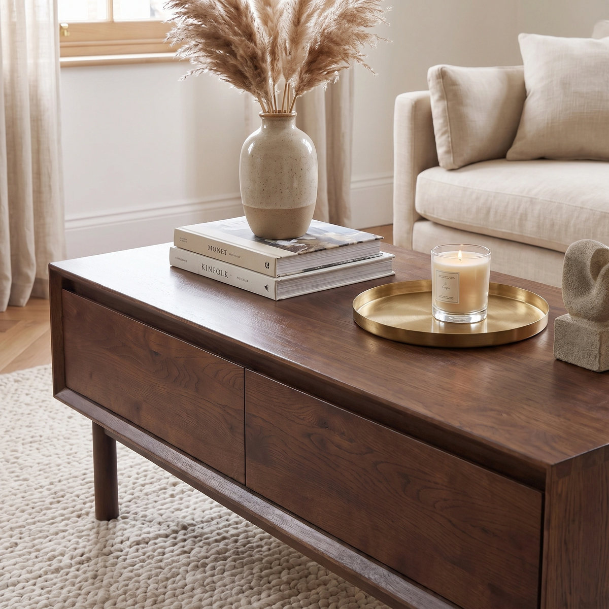 The Oslo Coffee Table with 4 Drawers in Walnut, styled with books, a vase of pampas grass, a candle, and a gold tray, creates an inviting centerpiece in this cozy living room.