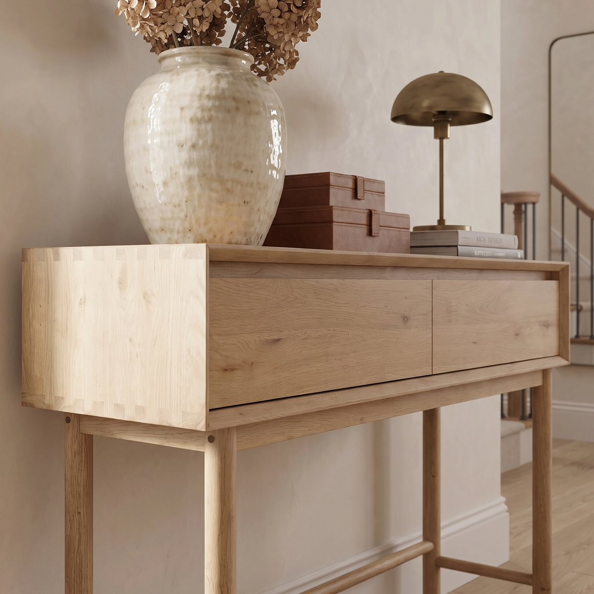 Embodying minimalist charm, the Oslo 2 Drawer Console Table in Light Oak showcases a ceramic vase, dried flowers, brown boxes, and a brass lamp arranged gracefully on its surface.