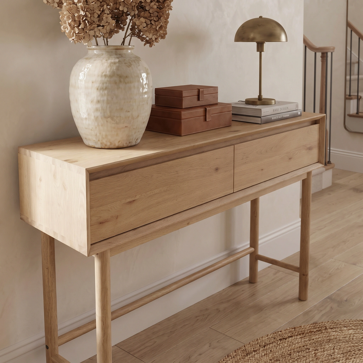 Oslo 2 Drawer Console Table in Light Oak styled with a vase, lamp, stacked books, and decorative boxes in a hallway.