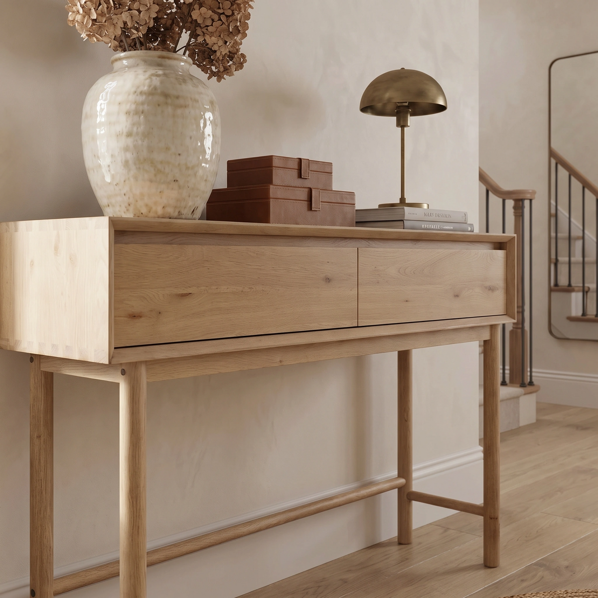 The Oslo 2 Drawer Console Table in Light Oak is beautifully styled with dried flowers, stacked boxes, books, and a brass lamp in a neutral-toned hallway.