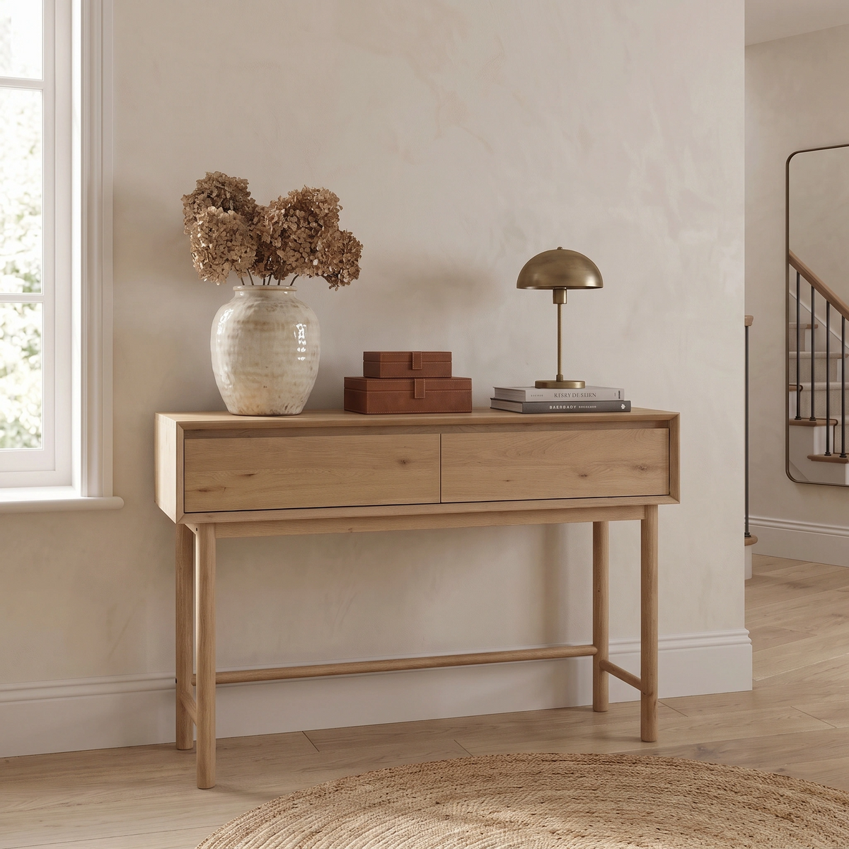 Oslo 2 Drawer Console Table in Light Oak, styled with a vase of dried flowers, boxes, and a lamp in a bright, modern room.