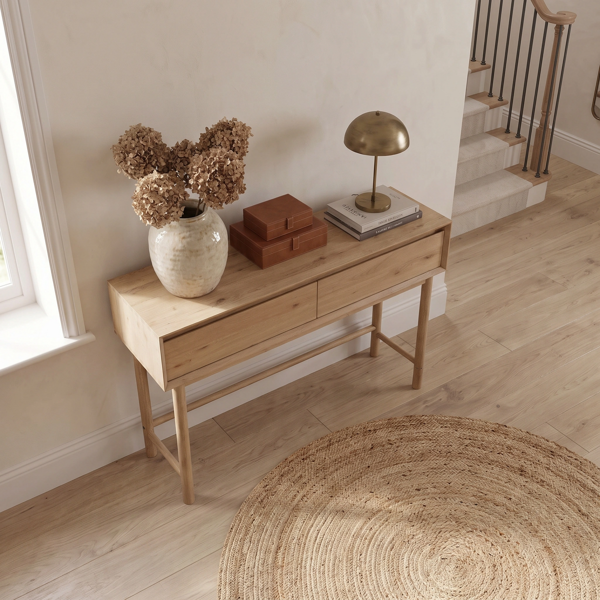 The Oslo 2 Drawer Console Table in Light Oak holds a vase of dried hydrangeas, two brown leather boxes, and a gold lamp near a staircase.