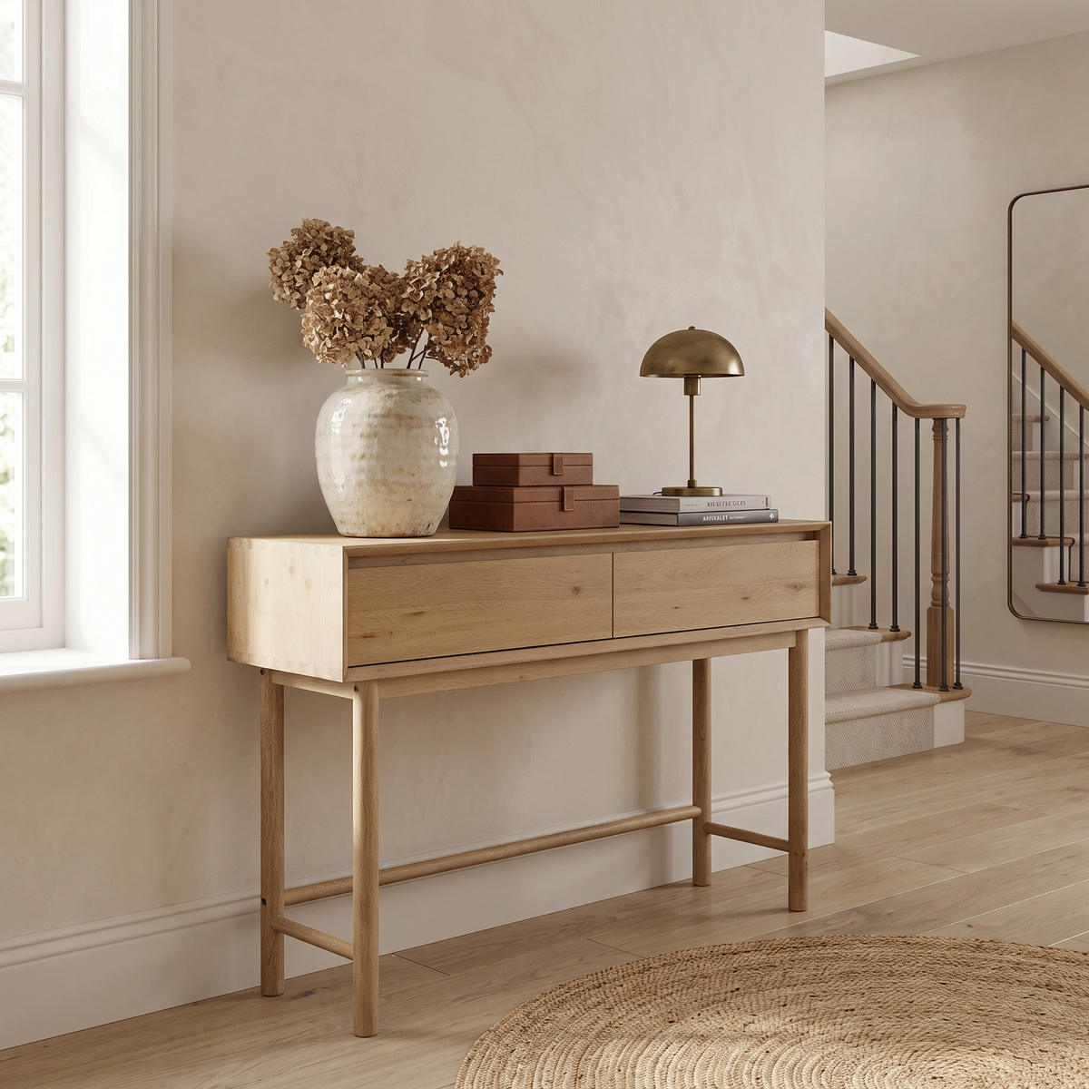 Oslo 2 Drawer Console Table in Light Oak styled with a vase of dried flowers, stacked books, and a brass lamp in a bright hallway.
