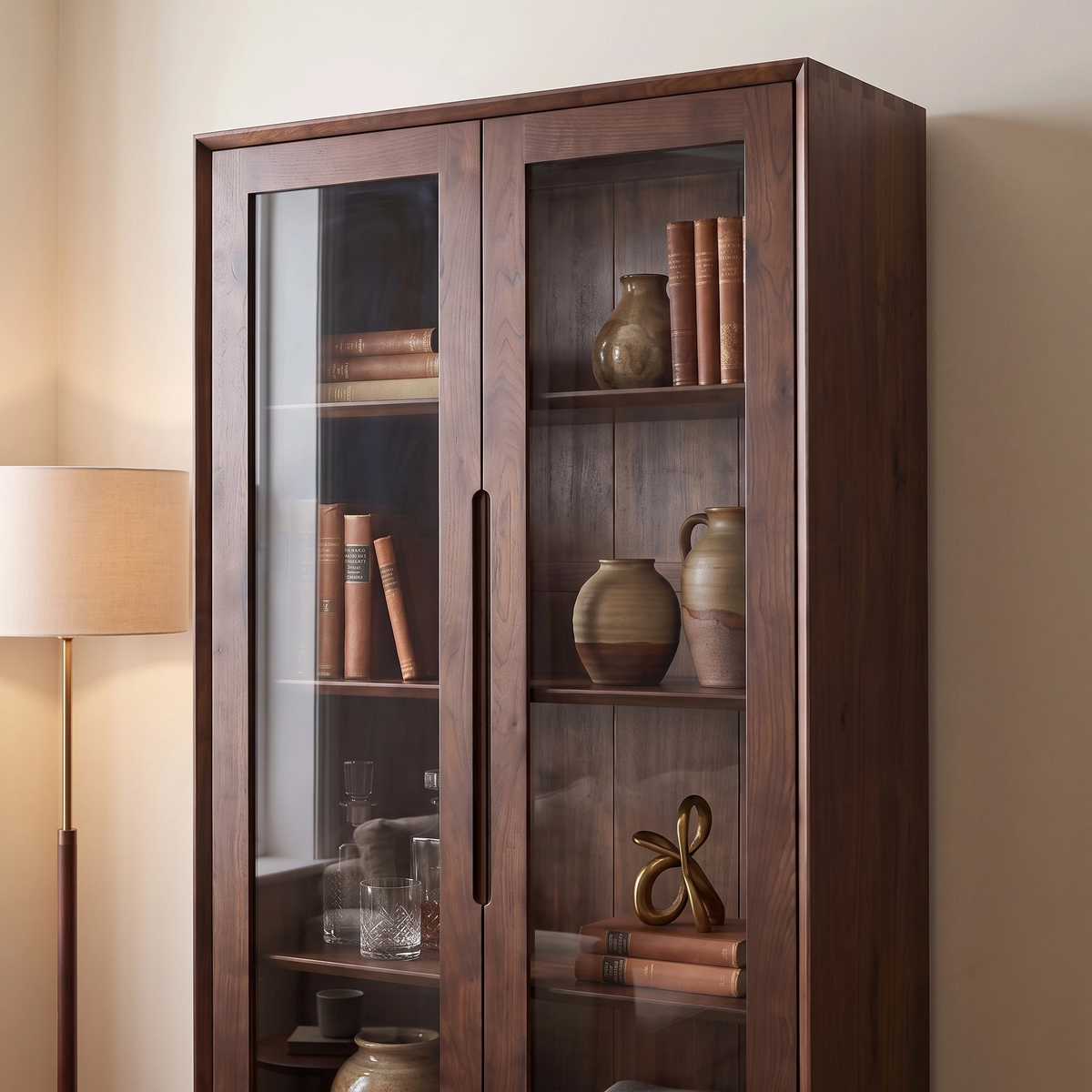 The Oslo Display Cabinet in Walnut, featuring glass doors, showcases books, pottery, and decor items next to a floor lamp.