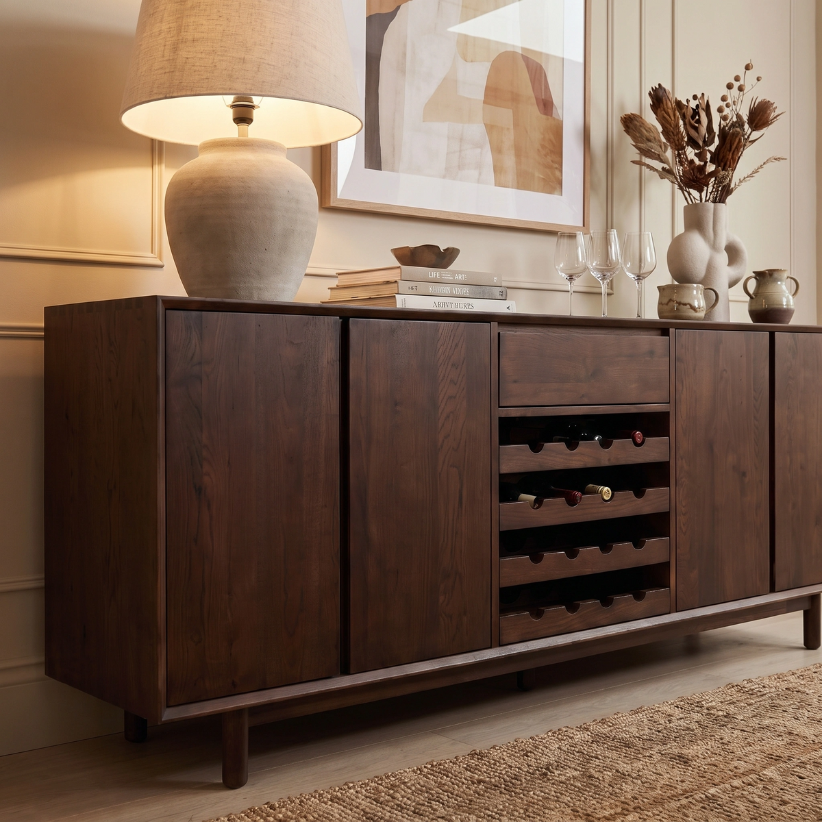 The Oslo Grand 4 Door Sideboard with Wine Rack in Walnut features a built-in wine rack and a spacious four-door design, complemented by a stylish lamp, books, glasses, and dried flowers in a cozy room.