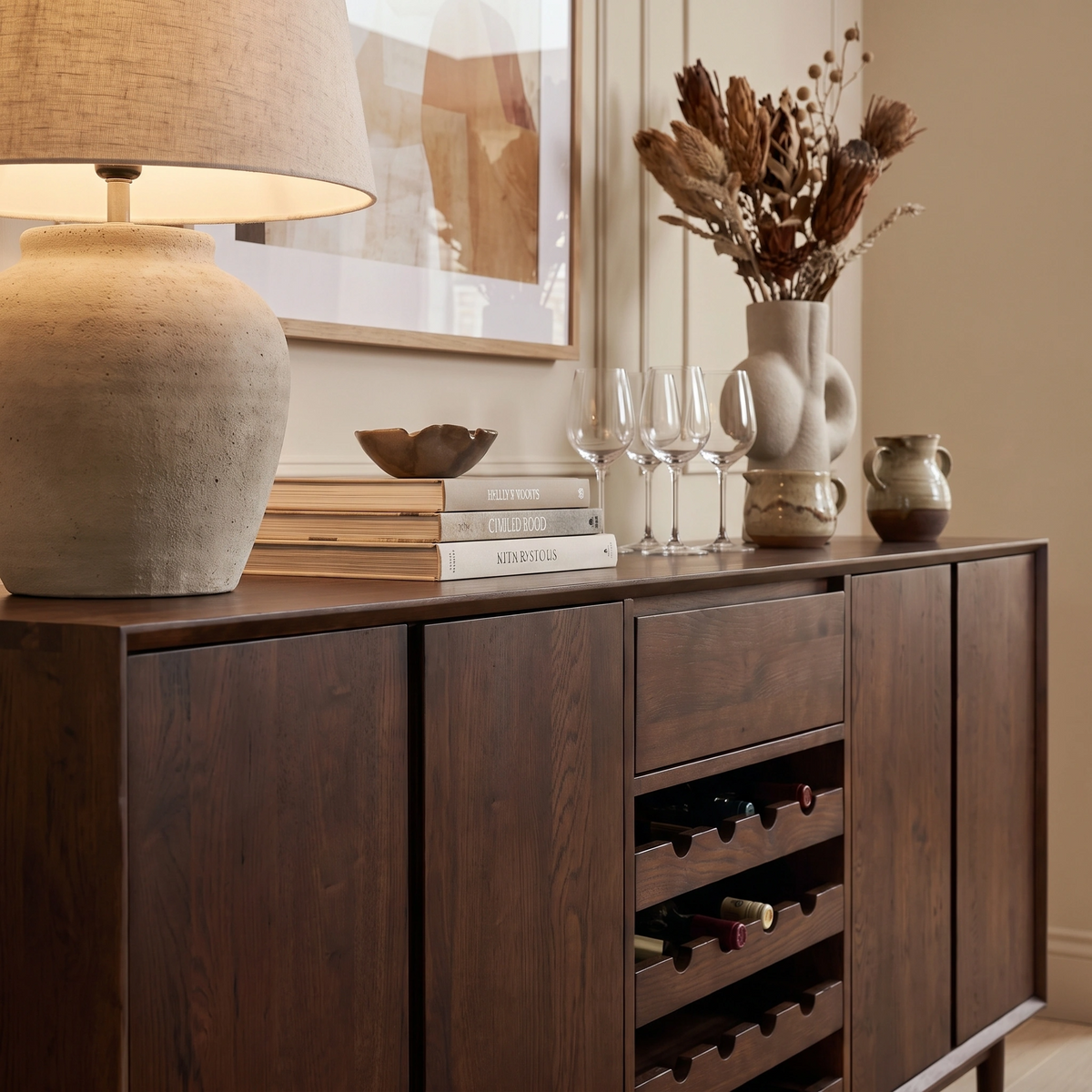 Oslo Grand 4 Door Sideboard with Wine Rack in Walnut, adorned with glassware, a lamp, dried flowers, and stacked books in a cozy room.