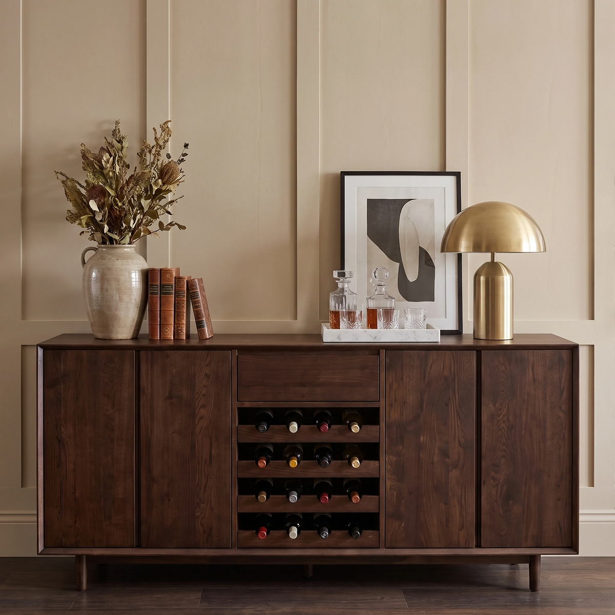 The Oslo Grand 4 Door Sideboard with Wine Rack in Walnut, elegantly topped with books, a vase of dried flowers, art, decanters, and a gold lamp—a sophisticated storage solution perfect for stylish display.