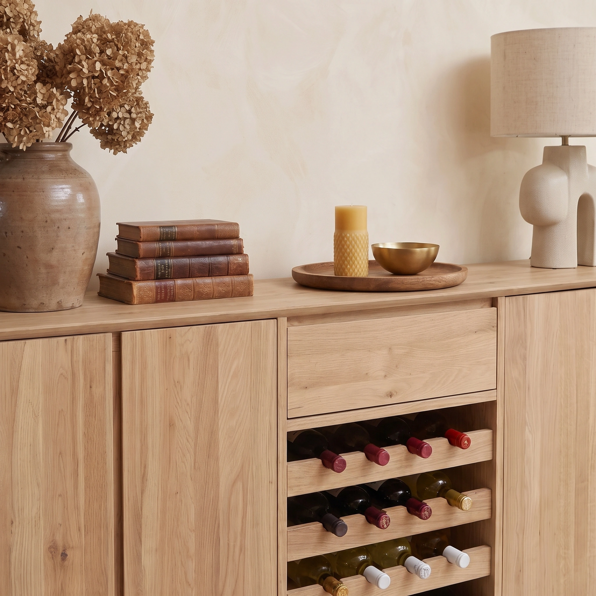 An Oslo Grand 4 Door Sideboard with Wine Rack in Light Oak is styled with a vase of dried flowers, vintage books, and a candle for an inviting touch, while its built-in wine rack is filled with wine bottles.