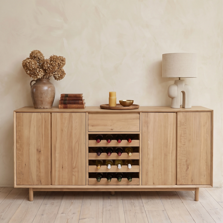 The Oslo Grand 4 Door Sideboard with Wine Rack in Light Oak is elegantly styled with a lamp, dried flowers, books, and decor, all displayed on a pale wood floor.