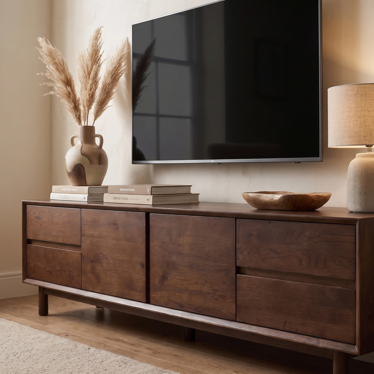 A stylish Oslo TV Unit in Walnut sits neatly under a wall-mounted TV, adorned with books, dried pampas grass in a vase, a lamp, and a wooden bowl, bringing warmth and elegance to any living space.