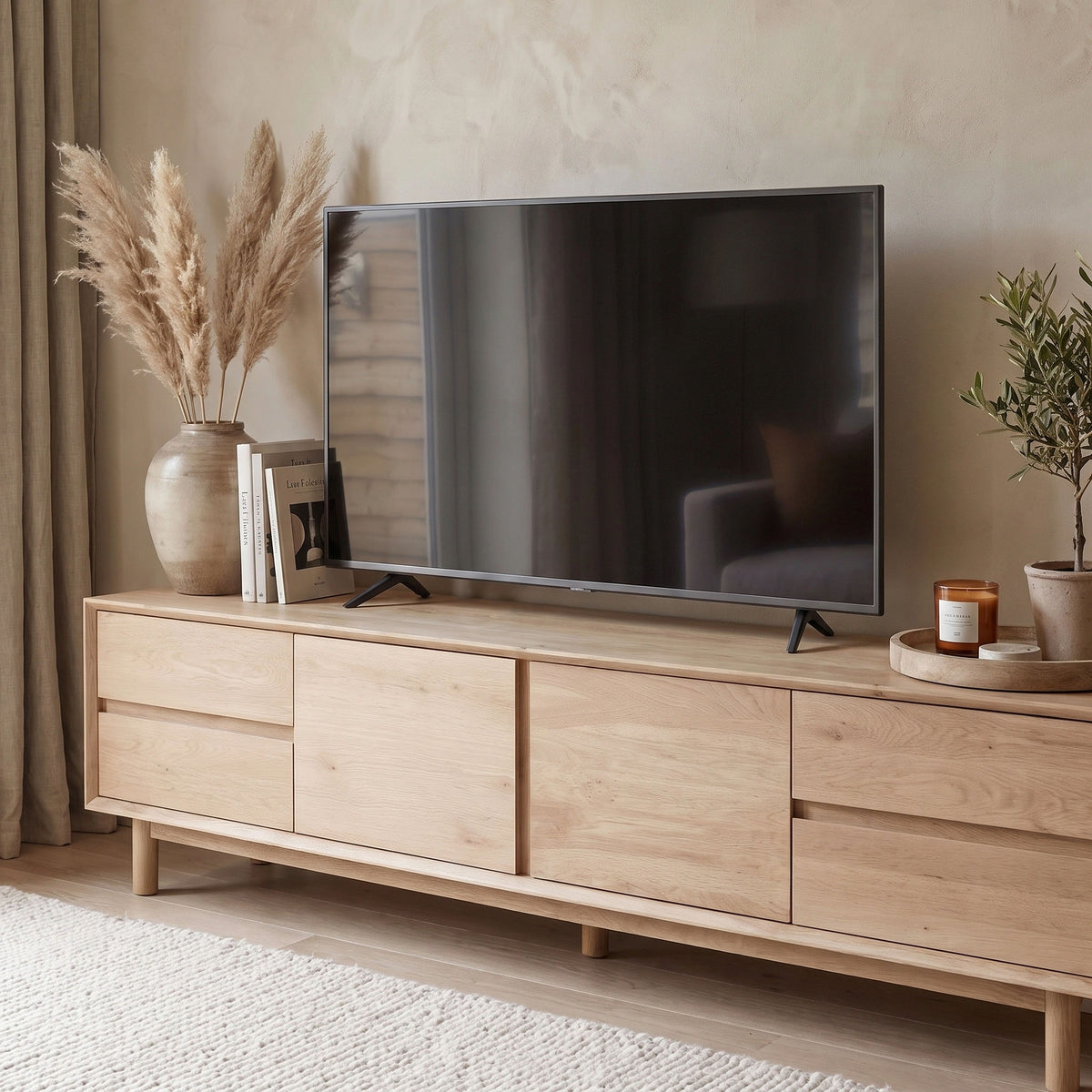 The Oslo TV Unit in Light Oak displays a flat-screen TV, complemented by vases, pampas grass, a candle, and a plant in a cozy living room decorated with modern furniture.