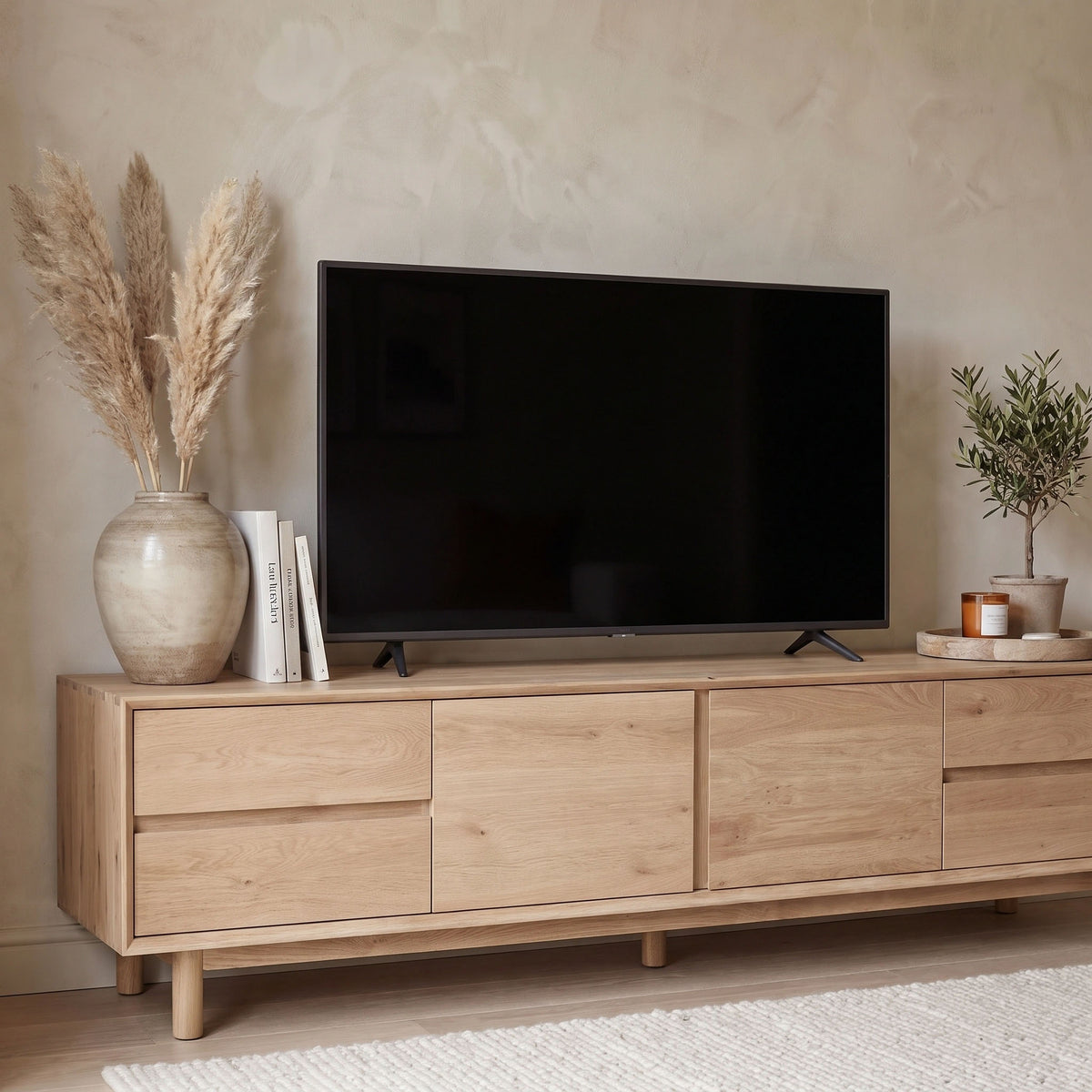 The Oslo TV Unit in Light Oak serves as a stylish modern media cabinet, displayed with books, a vase of pampas grass, a small plant, and a candle beside a flat-screen TV—an ideal addition to contemporary living room furniture.