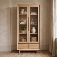 The Oslo Display Cabinet in Light Oak features elegant glass doors and displays ceramics, baskets, and a potted plant in a neutral-toned room.