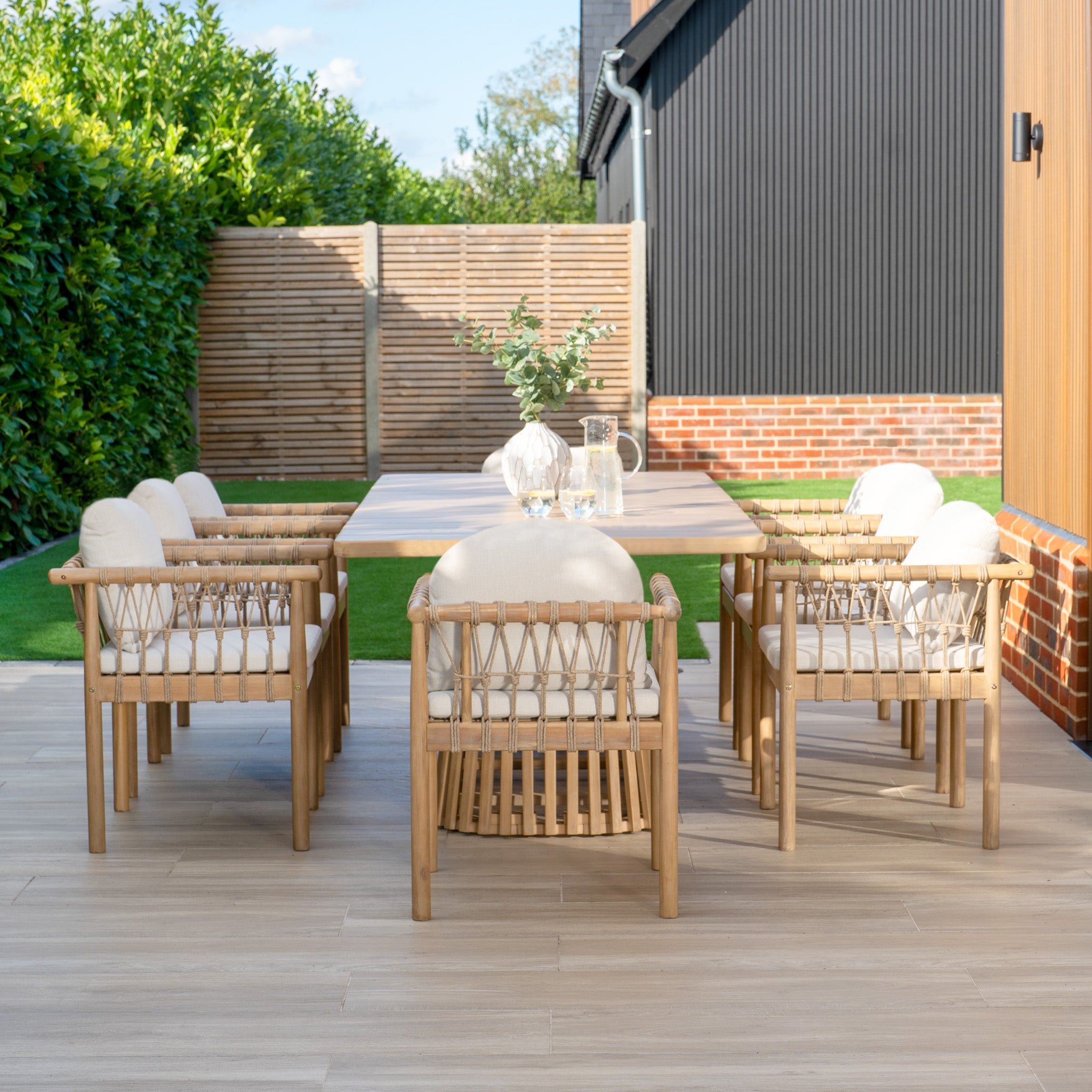 Enjoy outdoor dining with the Paia 8 Seat Rectangular Dining Set in Linen, complete with a sustainably crafted Acacia wood table and six cushioned chairs, set against a tranquil scene of a potted plant, wooden privacy fence, and lush grass.