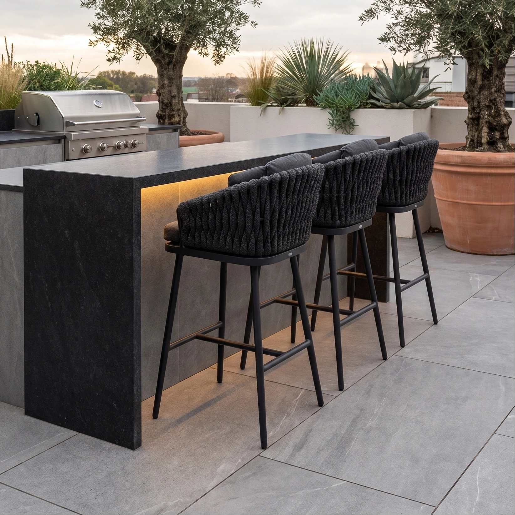 Modern outdoor kitchen with a black stone counter, four Palma Rope Bar Stools in Grey that are UV resistant, lush plants, and a grill.