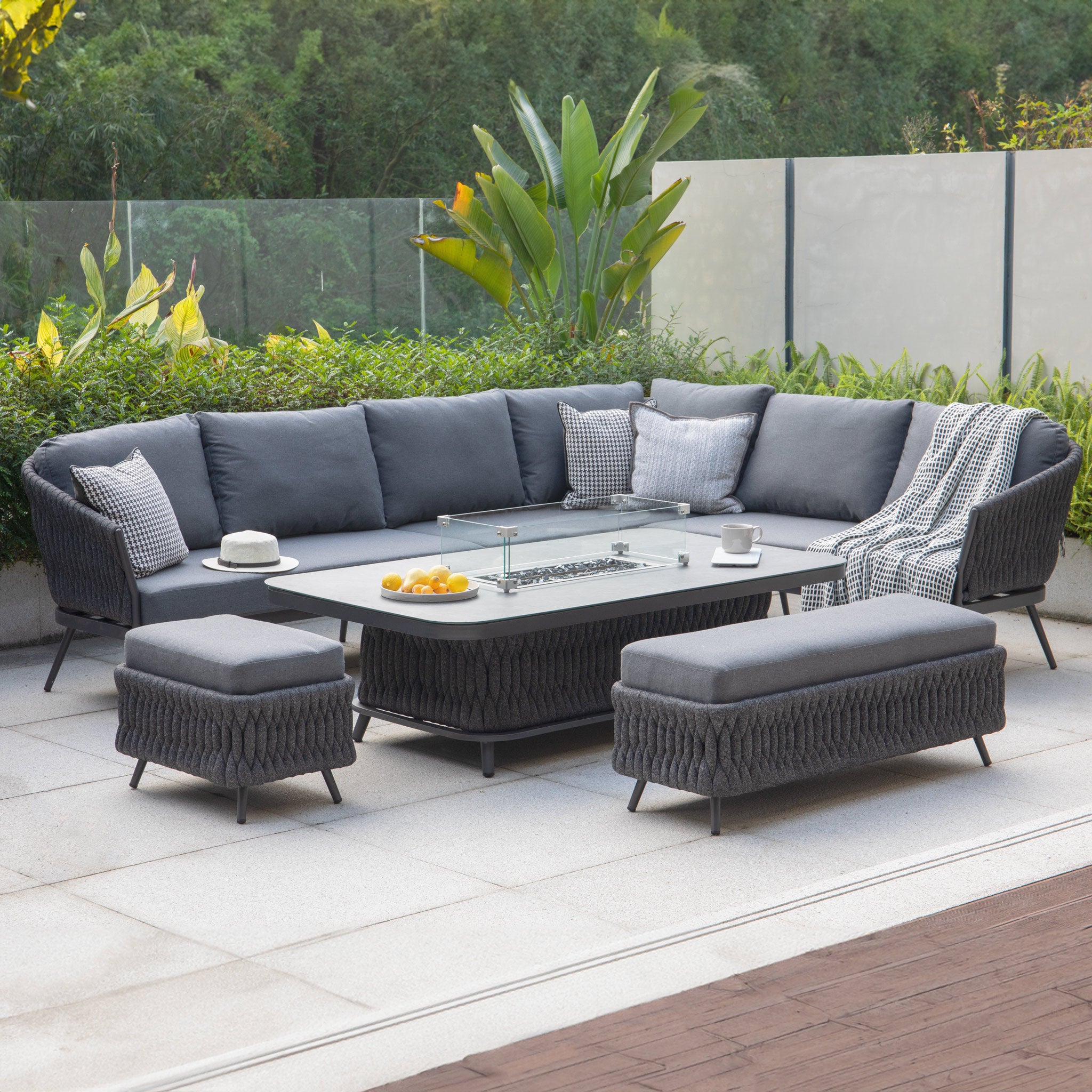 Outdoor patio furniture set featuring the stylish Palma Rope Rectangular Corner Dining Set with Rising Firepit Table in Grey (Right Hand), complete with a grey sectional sofa, fire pit table, stools, and lush greenery in the background. The water-repelling thick rope weave and aluminium frames ensure durability and comfort for any gathering.