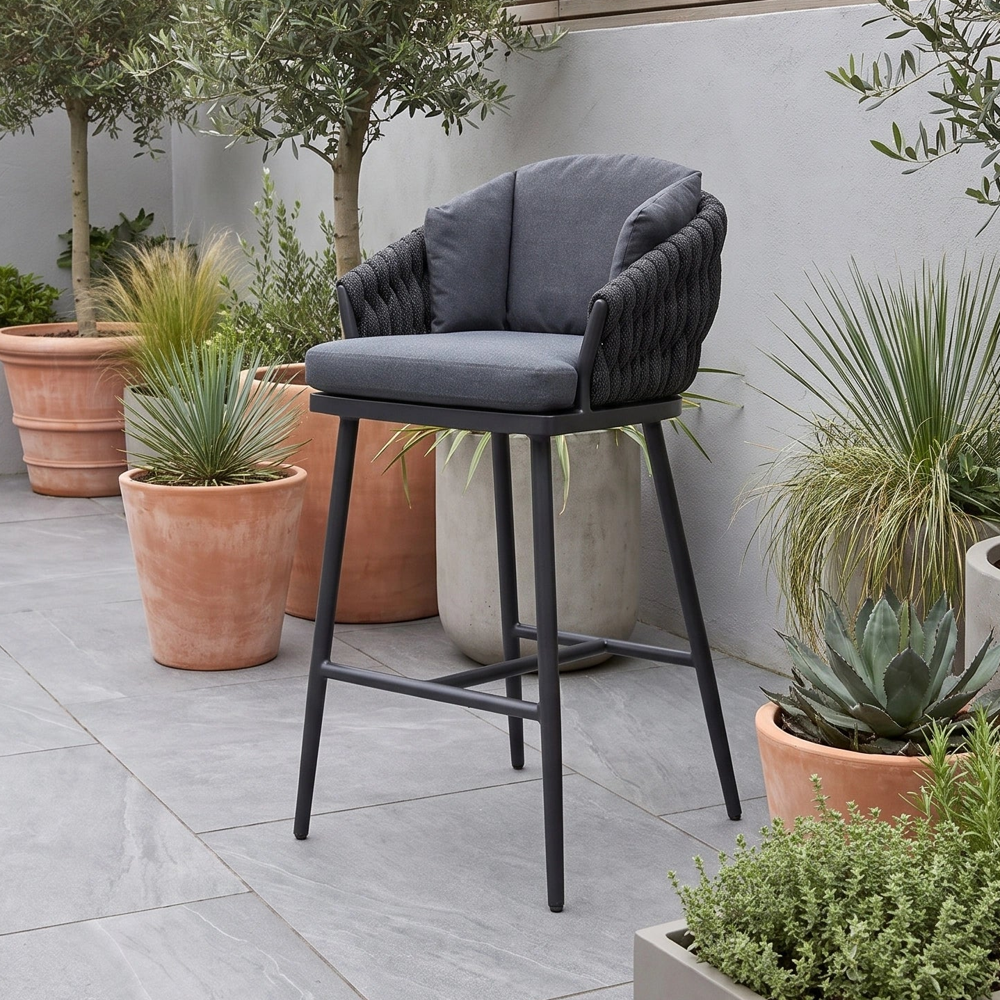 A Palma Rope Bar Stool in Grey sits on a patio surrounded by potted plants and greenery, offering both style and durability as a UV resistant bar stool for any outdoor setting.