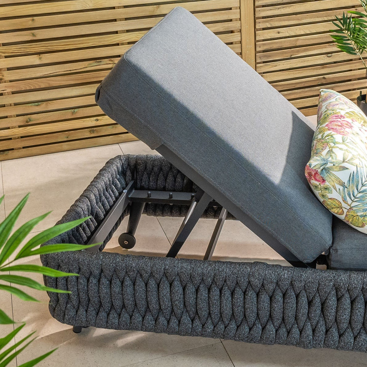 The Palma Rope Sun Lounger in Grey, featuring fade-resistant cushions with a floral pattern, is ideal as weatherproof garden furniture near wooden slats and lush green plants.